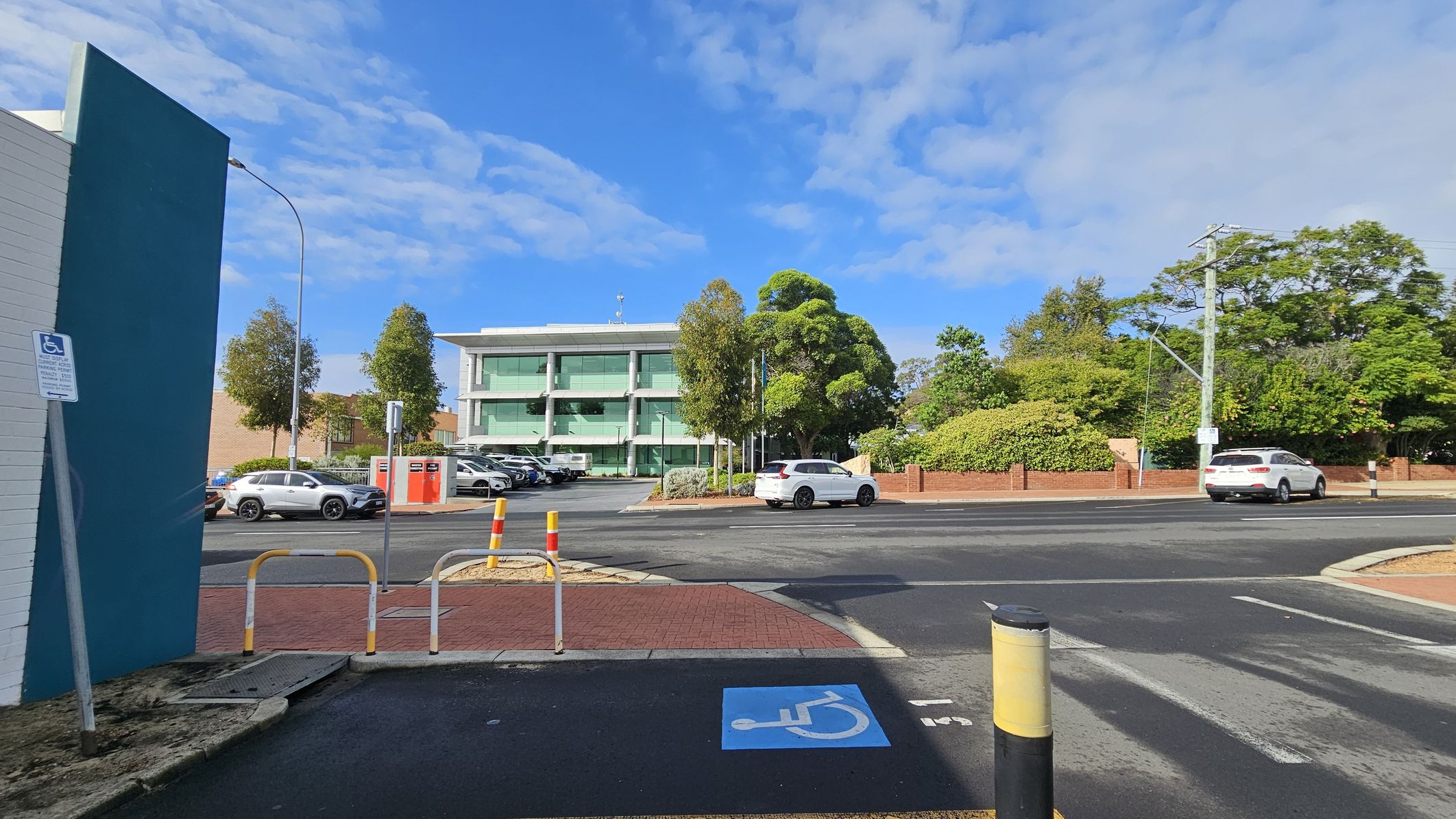A street scene with a three-storey building in the centre, featuring large glass windows. In front of the building, there are several parked cars and trees. On the left, a blue wall with a disabled parking sign is visible. The foreground shows a disabled parking space marked on the ground. On the right, a white car is driving past, with more trees and a brick wall in the background. The road is bordered by a red brick pavement. A street scene with a three-storey building in the centre, featuring large glass windows. In front of the building, there are several parked cars and trees. On the left, a blue wall with a disabled parking sign is visible. The foreground shows a disabled parking space marked on the ground. On the right, a white car is driving past, with more trees and a brick wall in the background. The road is bordered by a red brick pavement.
