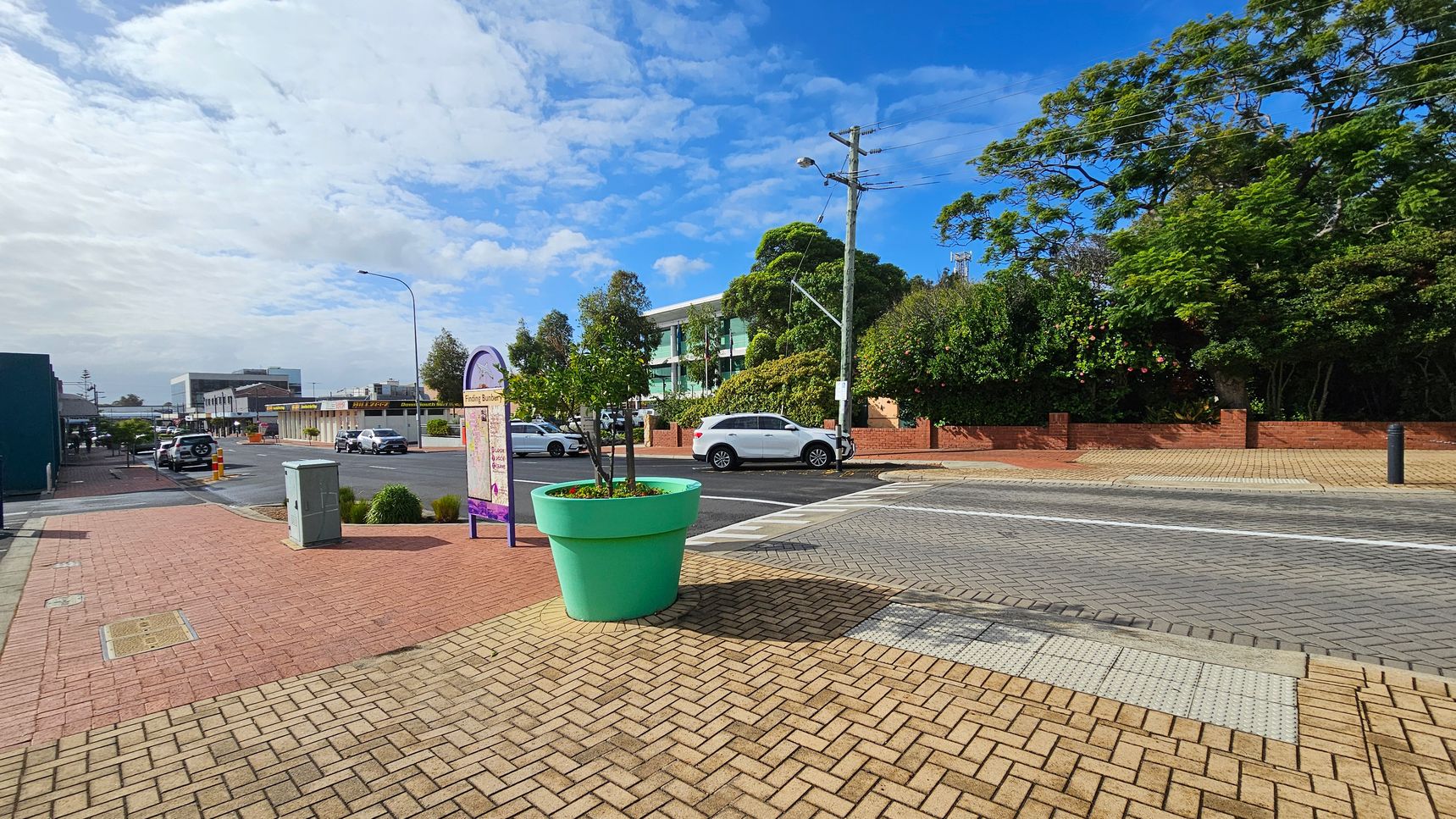A street scene with a large turquoise planter in the foreground, containing small trees. The pavement is made of light brown bricks. To the left, a sign with a map stands near a grey utility box. In the background, a white car is parked on the road, with a building partially obscured by trees. The right side features a brick wall and dense greenery. The sky is partly cloudy. A street scene with a large turquoise planter in the foreground, containing small trees. The pavement is made of light brown bricks. To the left, a sign with a map stands near a grey utility box. In the background, a white car is parked on the road, with a building partially obscured by trees. The right side features a brick wall and dense greenery. The sky is partly cloudy.