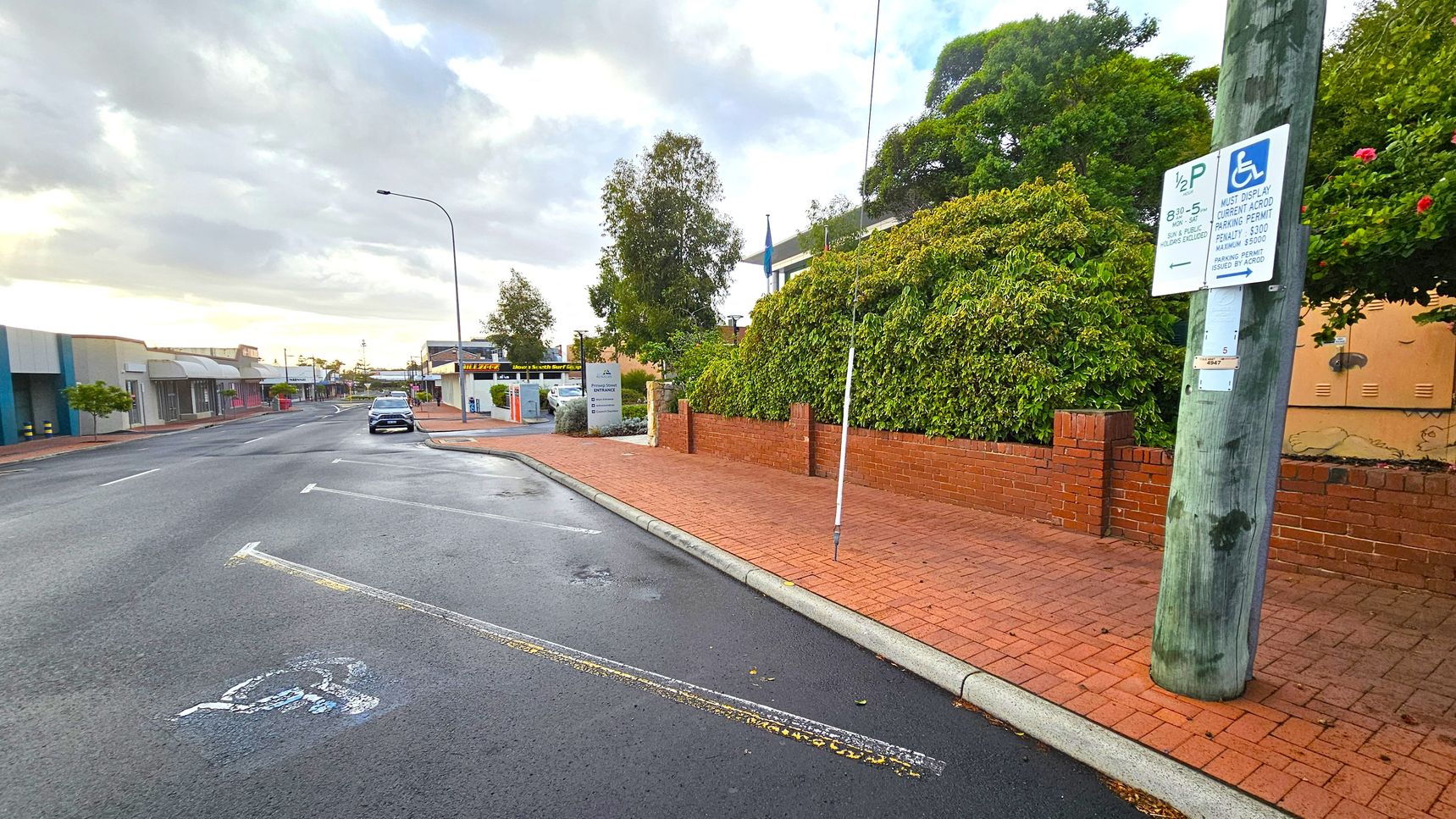 A street scene with a wet road and a single parked car on the left. The pavement is red brick, bordered by a low red brick wall with dense green foliage behind it. A wooden utility pole on the right has a sign indicating disabled parking. Buildings with shopfronts line the left side of the street, and a tall streetlamp is visible. The sky is overcast, and the area appears quiet. A street scene with a wet road and a single parked car on the left. The pavement is red brick, bordered by a low red brick wall with dense green foliage behind it. A wooden utility pole on the right has a sign indicating disabled parking. Buildings with shopfronts line the left side of the street, and a tall streetlamp is visible. The sky is overcast, and the area appears quiet.
