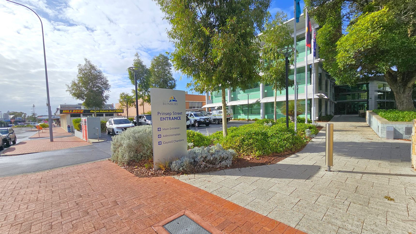 A paved walkway leads to a modern glass building on the right, partially obscured by trees and greenery. A sign in the foreground reads "Prinsep Street Entrance" with directions. On the left, a street with parked cars and a shop sign is visible. The pavement is a mix of red and grey bricks, and there are tall street lamps along the path. Flags are flying near the building entrance. A paved walkway leads to a modern glass building on the right, partially obscured by trees and greenery. A sign in the foreground reads "Prinsep Street Entrance" with directions. On the left, a street with parked cars and a shop sign is visible. The pavement is a mix of red and grey bricks, and there are tall street lamps along the path. Flags are flying near the building entrance.