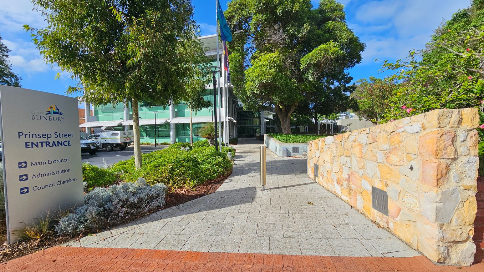 A paved walkway leads to a modern building with green-tinted glass windows in the background. On the left, a sign reads "City of Bunbury, Prinsep Street Entrance" with directions. A tree and a parked vehicle are nearby. On the right, a stone wall with plaques runs alongside the path, bordered by green shrubs and flowering plants. Several flags are visible near the building entrance. The area is well-landscaped with trees and bushes. A paved walkway leads to a modern building with green-tinted glass windows in the background. On the left, a sign reads "City of Bunbury, Prinsep Street Entrance" with directions. A tree and a parked vehicle are nearby. On the right, a stone wall with plaques runs alongside the path, bordered by green shrubs and flowering plants. Several flags are visible near the building entrance. The area is well-landscaped with trees and bushes.