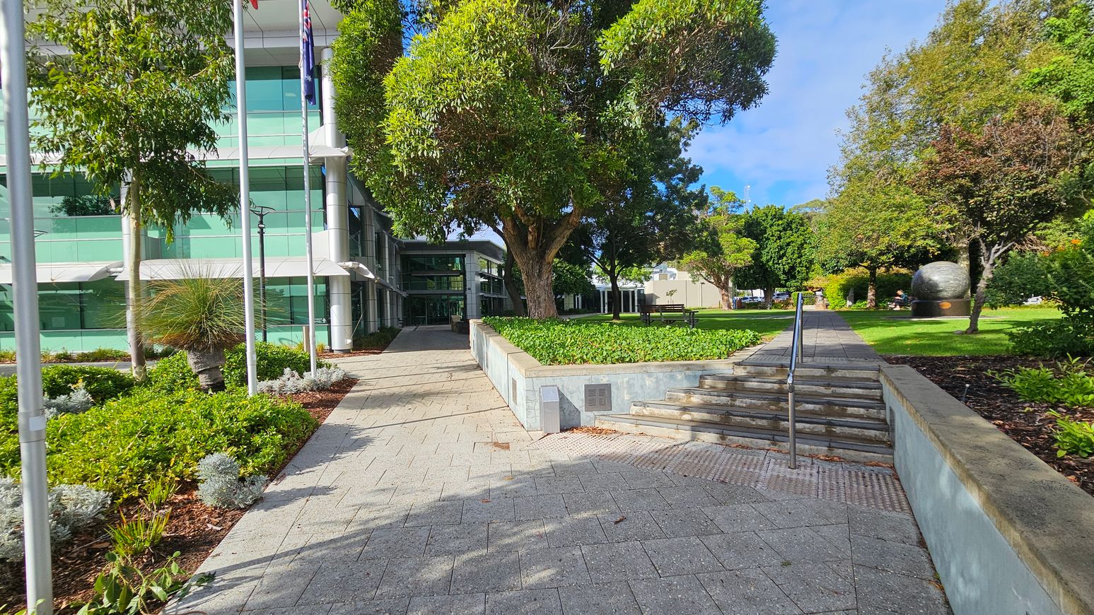 A modern building with large glass windows is on the left, partially obscured by trees and flagpoles. A paved walkway leads towards the entrance, flanked by landscaped greenery. In the centre, a large tree provides shade over a raised garden bed. On the right, a set of steps with a metal railing ascends to a grassy area with more trees and a large spherical sculpture. The ground is paved with grey tiles, and the area is well-maintained with various plants. A modern building with large glass windows is on the left, partially obscured by trees and flagpoles. A paved walkway leads towards the entrance, flanked by landscaped greenery. In the centre, a large tree provides shade over a raised garden bed. On the right, a set of steps with a metal railing ascends to a grassy area with more trees and a large spherical sculpture. The ground is paved with grey tiles, and the area is well-maintained with various plants.