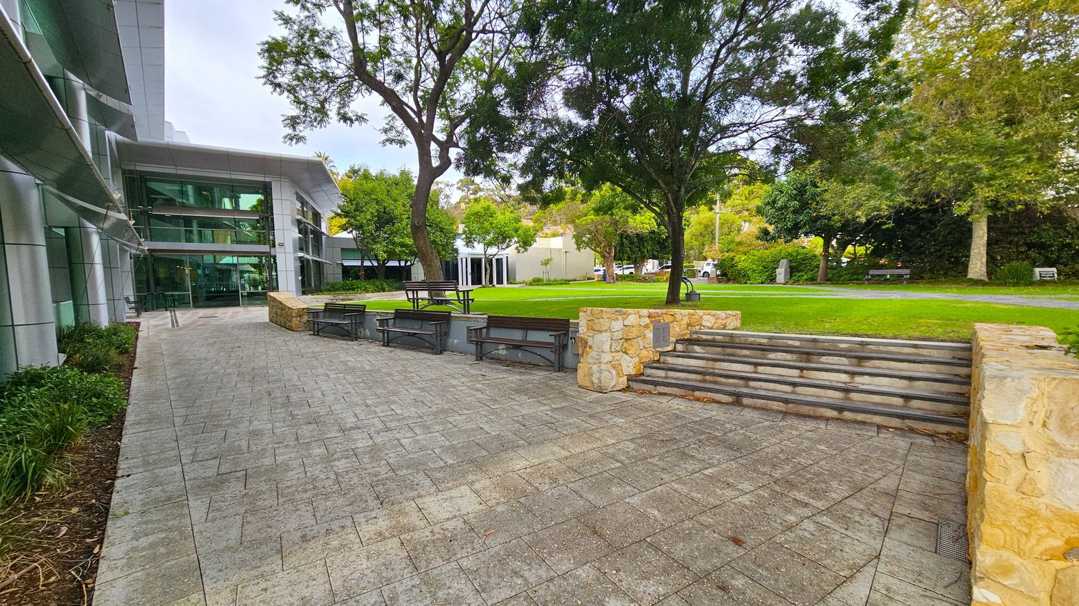 A modern building with large glass windows is on the left, adjacent to a paved walkway. In the centre, there are stone steps leading up to a grassy area with trees and several benches. The right side features more greenery and a stone wall. The background includes additional trees and a small structure, with a few parked vehicles visible. A modern building with large glass windows is on the left, adjacent to a paved walkway. In the centre, there are stone steps leading up to a grassy area with trees and several benches. The right side features more greenery and a stone wall. The background includes additional trees and a small structure, with a few parked vehicles visible.