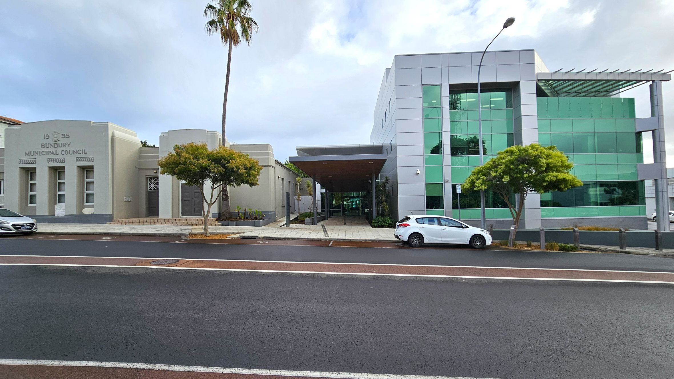 On the left, a beige building with "1935 Bunbury Municipal Council" inscribed above three windows. A palm tree and a smaller tree are in front. In the centre, a covered walkway leads to a modern building on the right, featuring green-tinted glass panels. A white car is parked in front of this building, and a small tree is nearby. The foreground shows a paved road with white and red markings. On the left, a beige building with "1935 Bunbury Municipal Council" inscribed above three windows. A palm tree and a smaller tree are in front. In the centre, a covered walkway leads to a modern building on the right, featuring green-tinted glass panels. A white car is parked in front of this building, and a small tree is nearby. The foreground shows a paved road with white and red markings.