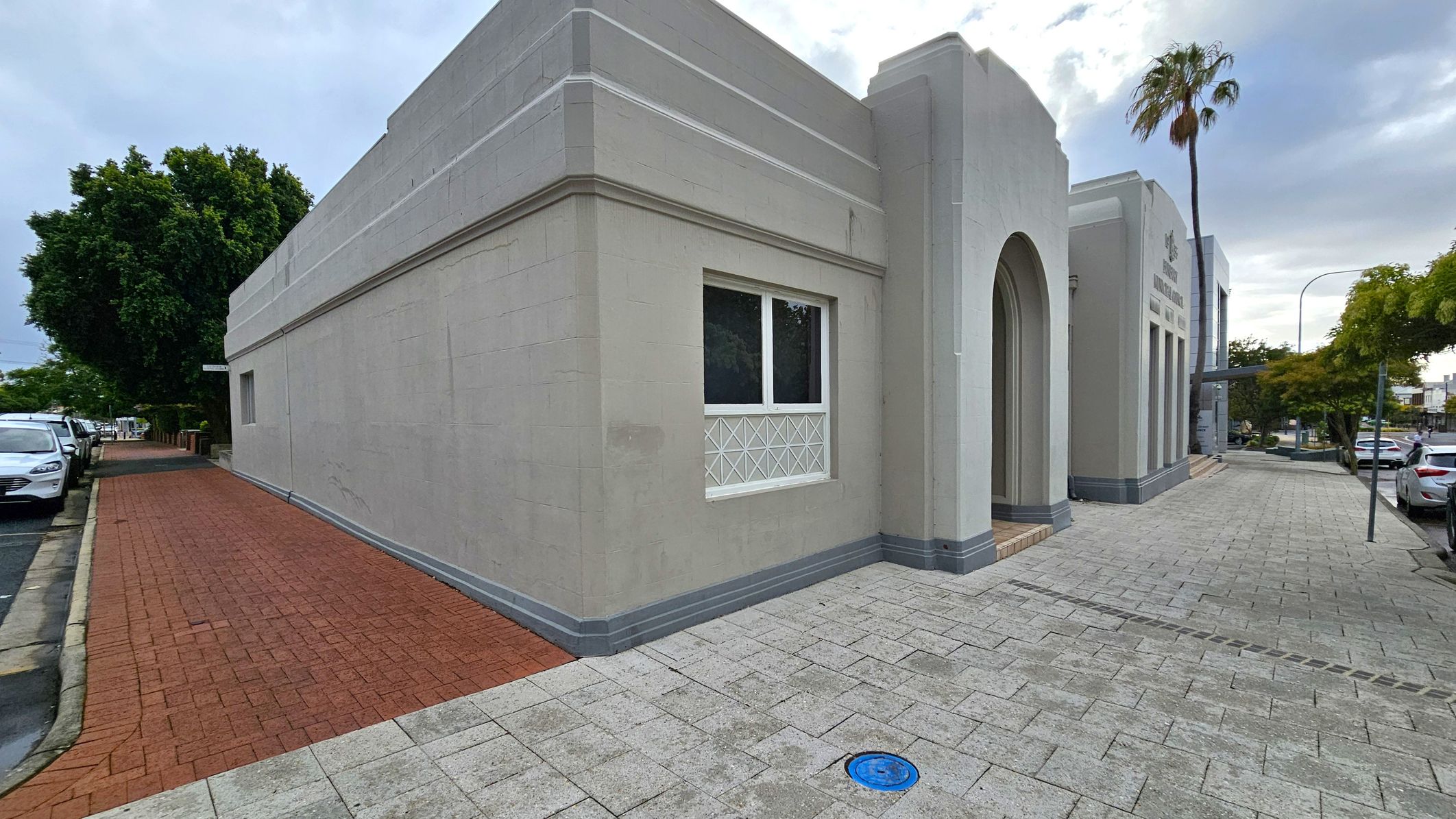A grey building with an arched entrance and rectangular windows is on the right. The pavement is made of light grey bricks, with a blue circular cover visible. A palm tree and other trees line the street, with parked cars along the kerb. On the left, a red brick path runs alongside the building, leading to more parked cars and greenery. A grey building with an arched entrance and rectangular windows is on the right. The pavement is made of light grey bricks, with a blue circular cover visible. A palm tree and other trees line the street, with parked cars along the kerb. On the left, a red brick path runs alongside the building, leading to more parked cars and greenery.