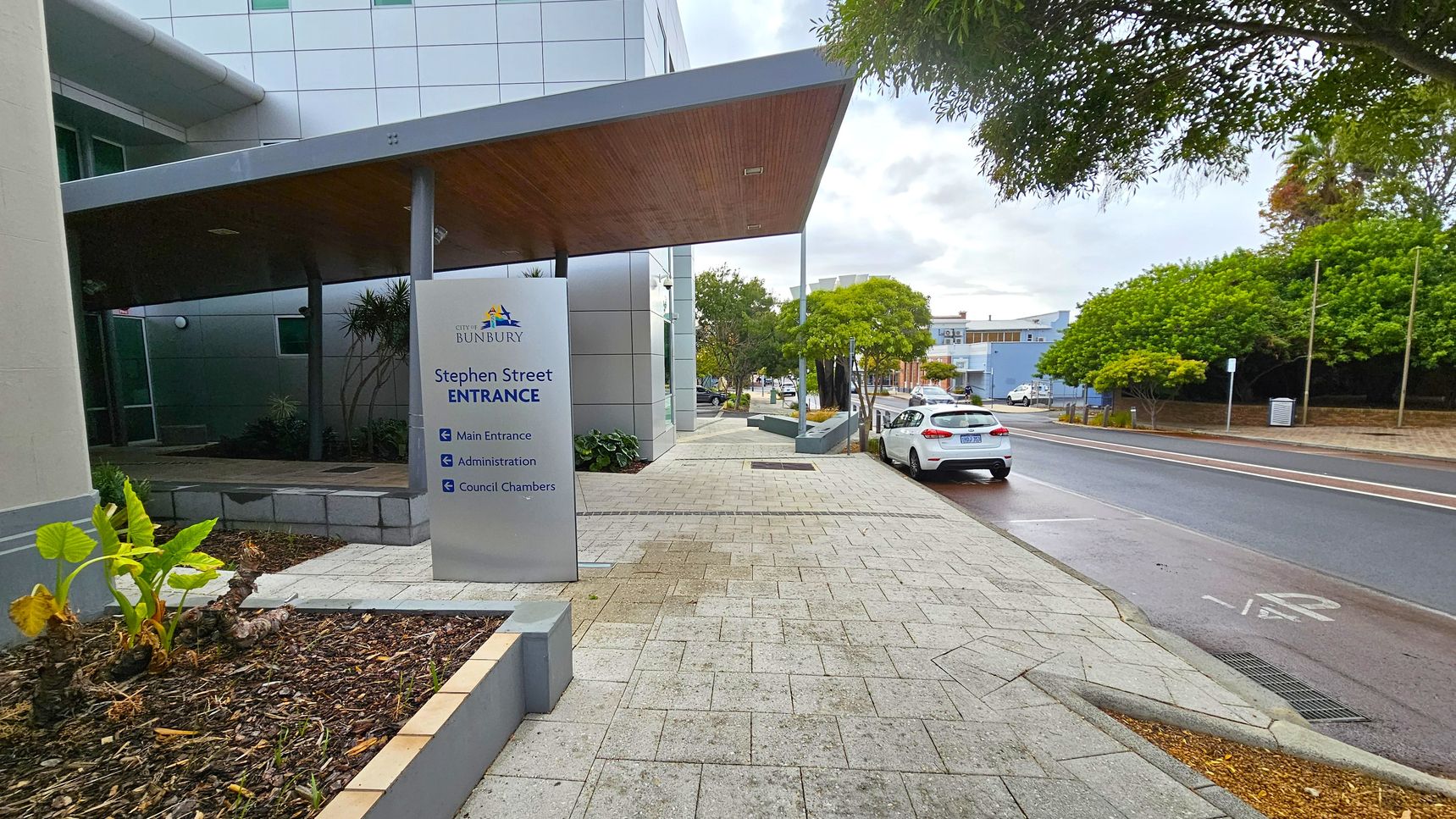 A paved walkway leads to a building with a sign reading "City of Bunbury, Stephen Street Entrance" on the left. The sign directs to the main entrance, administration, and council chambers. A modern building with a slanted roof is adjacent. On the right, a street with a parked white car and lush green trees lines the road. The pavement is light grey with a subtle pattern, and the area is well-maintained with small plants in a bed near the sign. A paved walkway leads to a building with a sign reading "City of Bunbury, Stephen Street Entrance" on the left. The sign directs to the main entrance, administration, and council chambers. A modern building with a slanted roof is adjacent. On the right, a street with a parked white car and lush green trees lines the road. The pavement is light grey with a subtle pattern, and the area is well-maintained with small plants in a bed near the sign.
