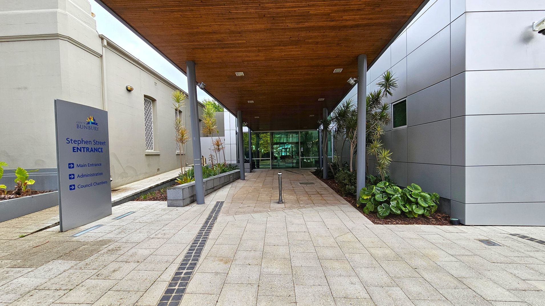A covered walkway leads to a glass entrance. On the left, a grey building with barred windows and a sign reading "Stephen Street Entrance" with directions to the main entrance, administration, and council chambers. On the right, a modern building with grey panels and plants along the side. The ceiling is wooden, and the floor is paved with light-coloured tiles. A covered walkway leads to a glass entrance. On the left, a grey building with barred windows and a sign reading "Stephen Street Entrance" with directions to the main entrance, administration, and council chambers. On the right, a modern building with grey panels and plants along the side. The ceiling is wooden, and the floor is paved with light-coloured tiles.