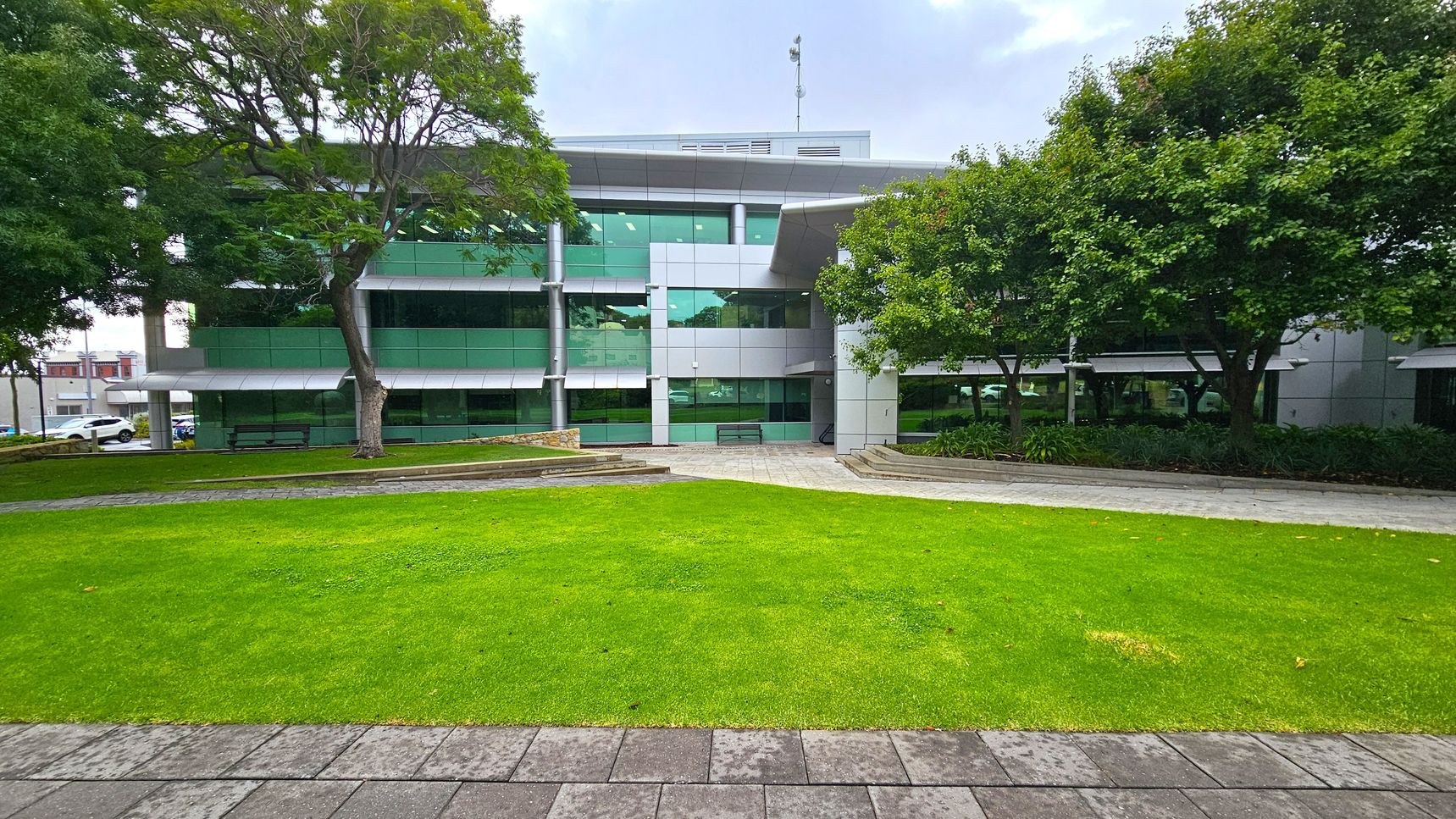 A modern building with large green-tinted windows and a grey facade is in the background. In the foreground, there is a well-maintained lawn bordered by a paved walkway. Trees with dense foliage are on both sides, providing shade. Benches are placed near the building, and there are landscaped areas with plants. The scene is calm and organised, with a mix of natural and architectural elements. A modern building with large green-tinted windows and a grey facade is in the background. In the foreground, there is a well-maintained lawn bordered by a paved walkway. Trees with dense foliage are on both sides, providing shade. Benches are placed near the building, and there are landscaped areas with plants. The scene is calm and organised, with a mix of natural and architectural elements.
