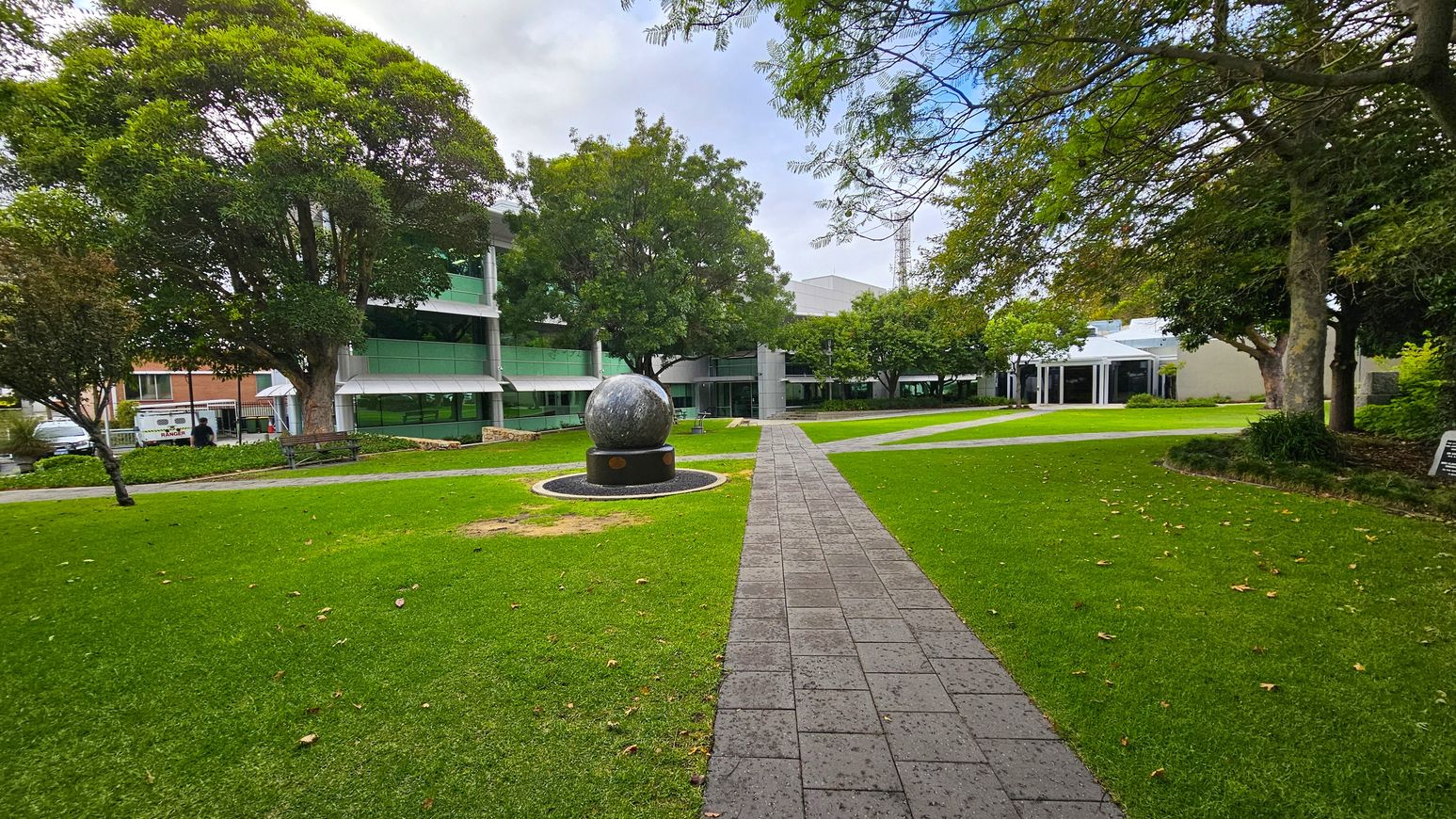 A paved path leads through a grassy area towards a large spherical sculpture on a pedestal in the centre. Surrounding the sculpture are well-maintained lawns with scattered fallen leaves. On the left, a large tree provides shade near a modern building with green-tinted windows. On the right, more trees and another building with a white facade are visible. The background features additional trees and structures, creating a serene park-like setting. A paved path leads through a grassy area towards a large spherical sculpture on a pedestal in the centre. Surrounding the sculpture are well-maintained lawns with scattered fallen leaves. On the left, a large tree provides shade near a modern building with green-tinted windows. On the right, more trees and another building with a white facade are visible. The background features additional trees and structures, creating a serene park-like setting.