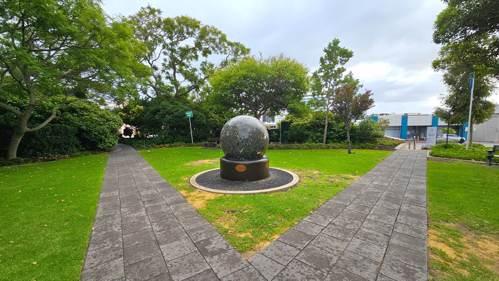 A spherical stone sculpture is centred on a circular base in a small park. Two paved paths intersect at the sculpture, forming a V-shape. Lush green grass surrounds the paths, with trees and bushes lining the edges. In the background, a modern building with blue accents is visible. A bench is partially visible to the left, and a signpost is near the centre. The sky is overcast. A spherical stone sculpture is centred on a circular base in a small park. Two paved paths intersect at the sculpture, forming a V-shape. Lush green grass surrounds the paths, with trees and bushes lining the edges. In the background, a modern building with blue accents is visible. A bench is partially visible to the left, and a signpost is near the centre. The sky is overcast.