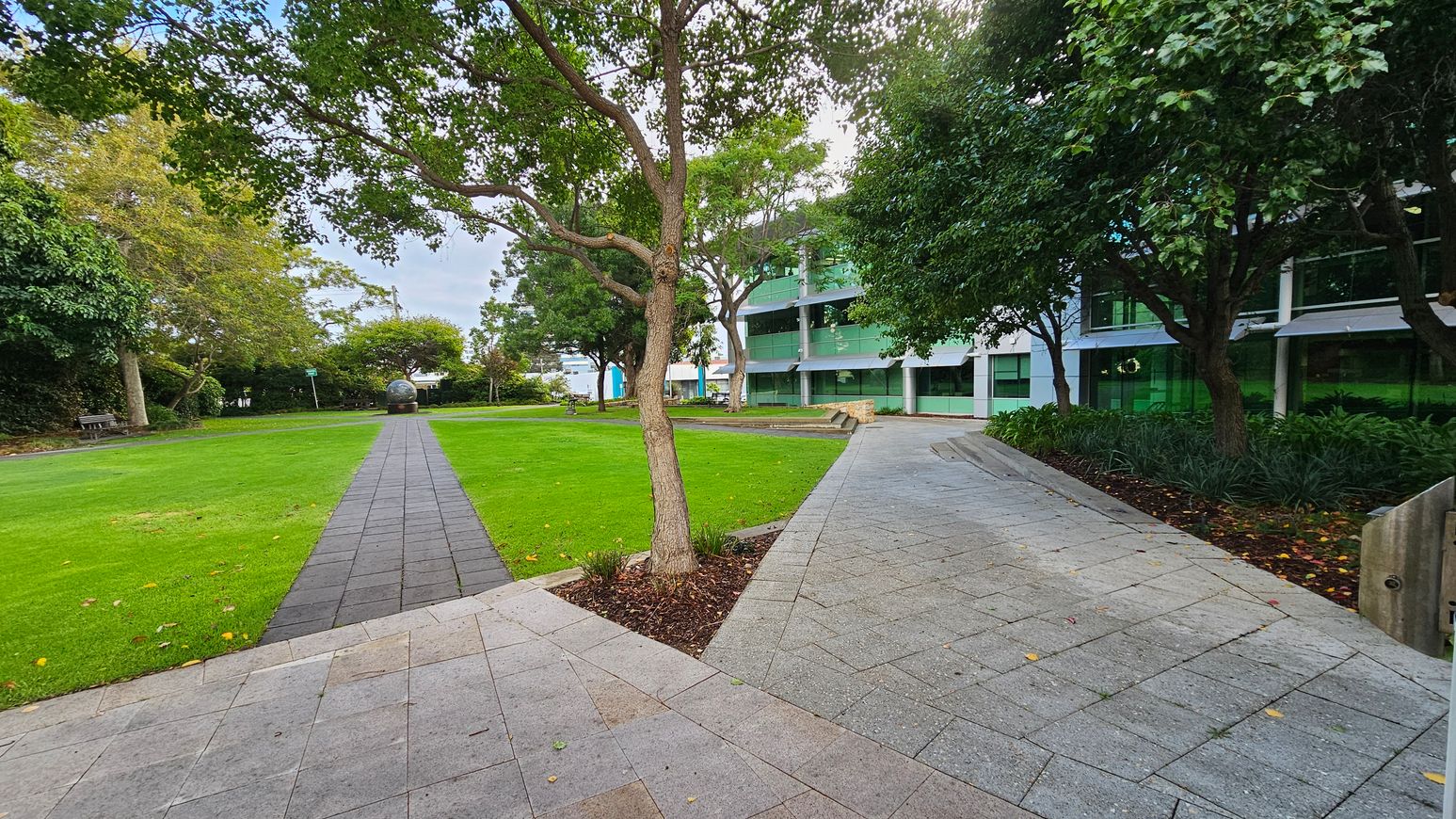 A paved walkway runs through a grassy area with trees on both sides. On the left, a bench is partially visible under the trees. In the centre, a spherical sculpture is situated at the end of the path. On the right, a modern building with large green-tinted windows is partially obscured by trees and shrubs. The ground is covered with grey paving stones, and the area is surrounded by lush greenery. A paved walkway runs through a grassy area with trees on both sides. On the left, a bench is partially visible under the trees. In the centre, a spherical sculpture is situated at the end of the path. On the right, a modern building with large green-tinted windows is partially obscured by trees and shrubs. The ground is covered with grey paving stones, and the area is surrounded by lush greenery.