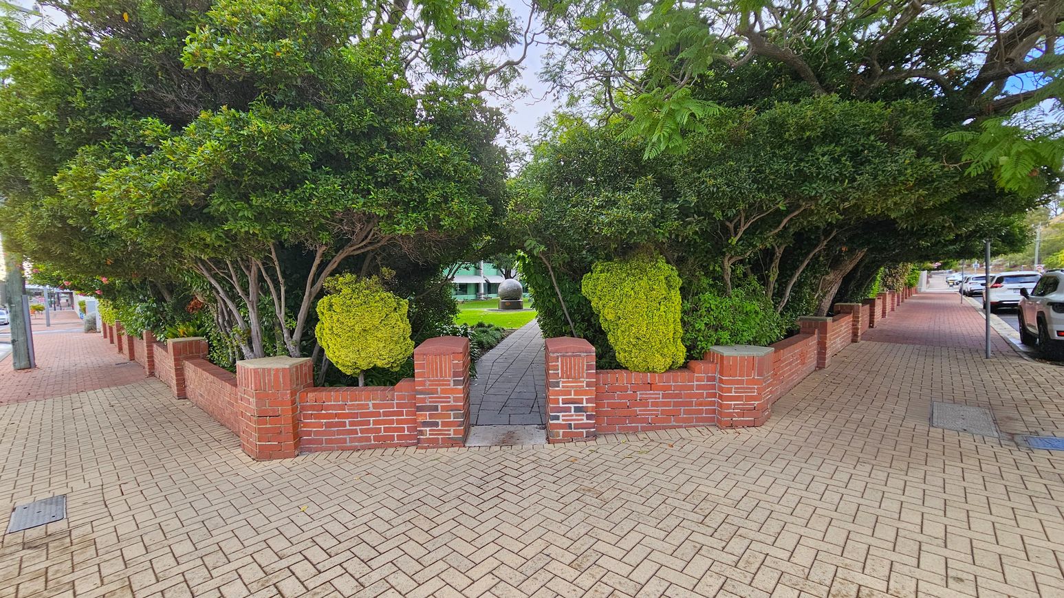A brick pathway leads through a small entrance flanked by red brick walls and dense green foliage. The path continues under an arch formed by trees and shrubs, leading to a grassy area with a large spherical sculpture in the distance. The foreground features a herringbone-patterned brick pavement, with parked cars and a street visible on the right. The left side shows more brick walls and trees lining the pavement. A brick pathway leads through a small entrance flanked by red brick walls and dense green foliage. The path continues under an arch formed by trees and shrubs, leading to a grassy area with a large spherical sculpture in the distance. The foreground features a herringbone-patterned brick pavement, with parked cars and a street visible on the right. The left side shows more brick walls and trees lining the pavement.