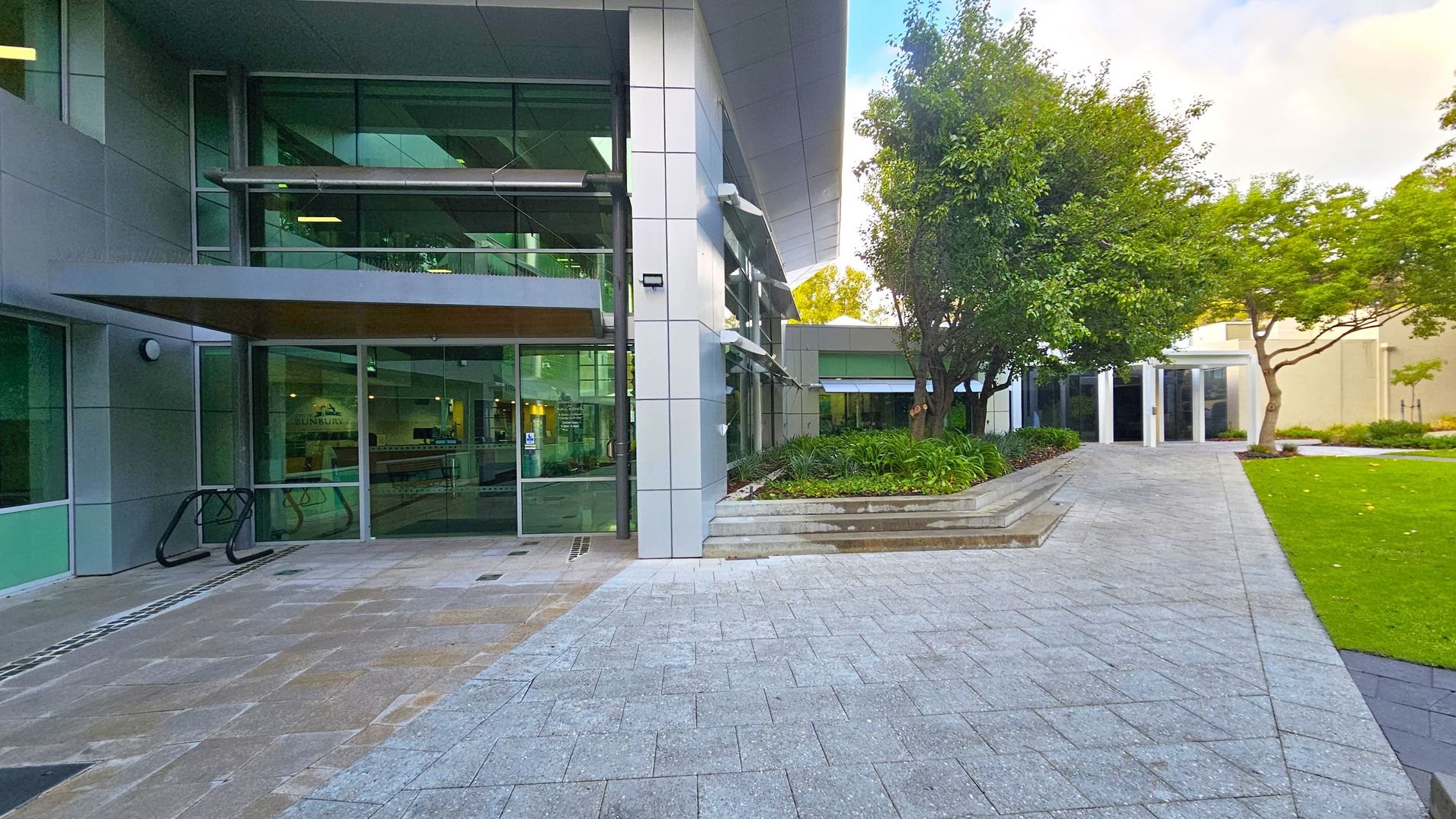 On the left, a modern building with large glass windows and a metal awning. A bike rack is visible near the entrance. In the centre, a paved walkway leads towards the building. On the right, a landscaped area with green grass, trees, and shrubs. A path extends towards another building entrance in the background. The scene is well-maintained and orderly. On the left, a modern building with large glass windows and a metal awning. A bike rack is visible near the entrance. In the centre, a paved walkway leads towards the building. On the right, a landscaped area with green grass, trees, and shrubs. A path extends towards another building entrance in the background. The scene is well-maintained and orderly.