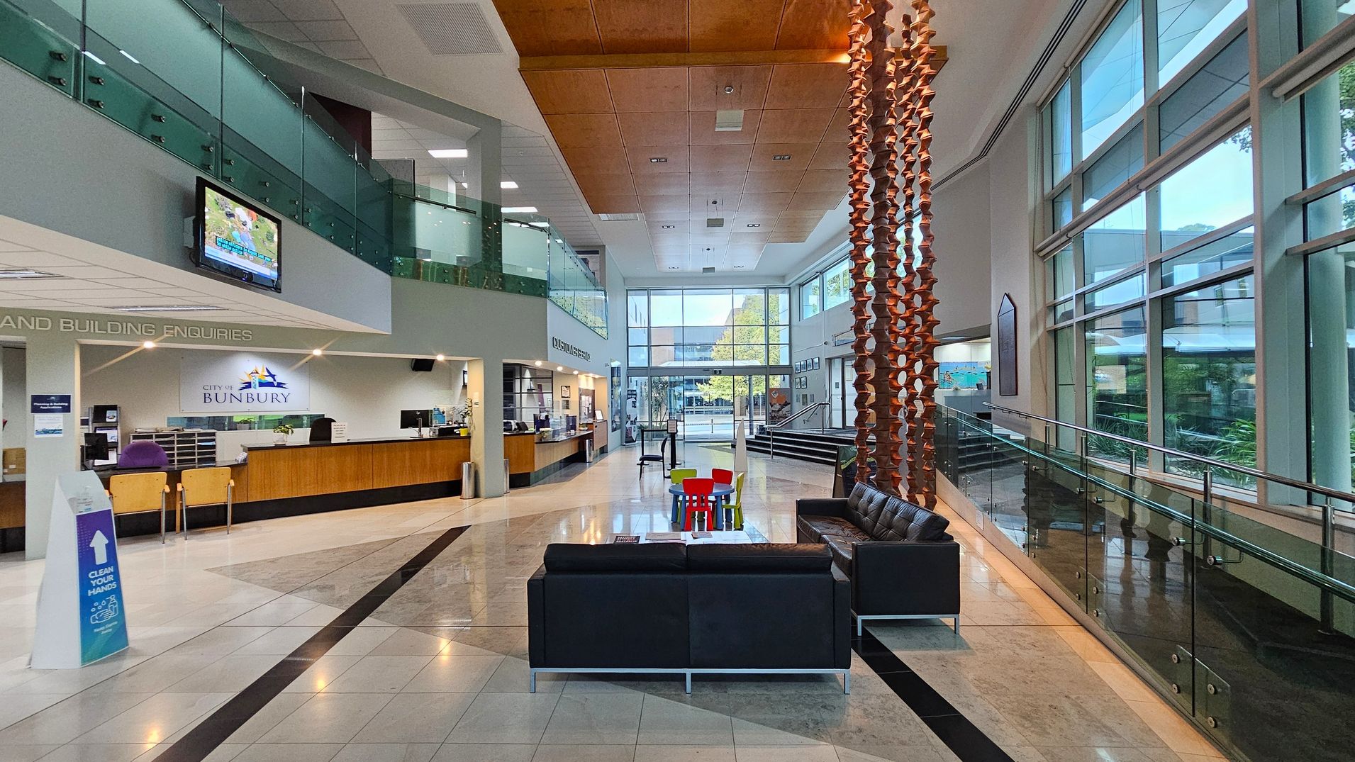 A spacious, modern lobby with a high ceiling and large windows on the right, allowing natural light to flood in. On the left, there's a reception desk with a sign reading "City of Bunbury" and a television mounted above. In the centre, a seating area with black sofas and a small table with colourful chairs. A decorative wooden sculpture hangs from the ceiling. The floor is polished with a geometric pattern, and a glass railing lines the right side. A spacious, modern lobby with a high ceiling and large windows on the right, allowing natural light to flood in. On the left, there's a reception desk with a sign reading "City of Bunbury" and a television mounted above. In the centre, a seating area with black sofas and a small table with colourful chairs. A decorative wooden sculpture hangs from the ceiling. The floor is polished with a geometric pattern, and a glass railing lines the right side.