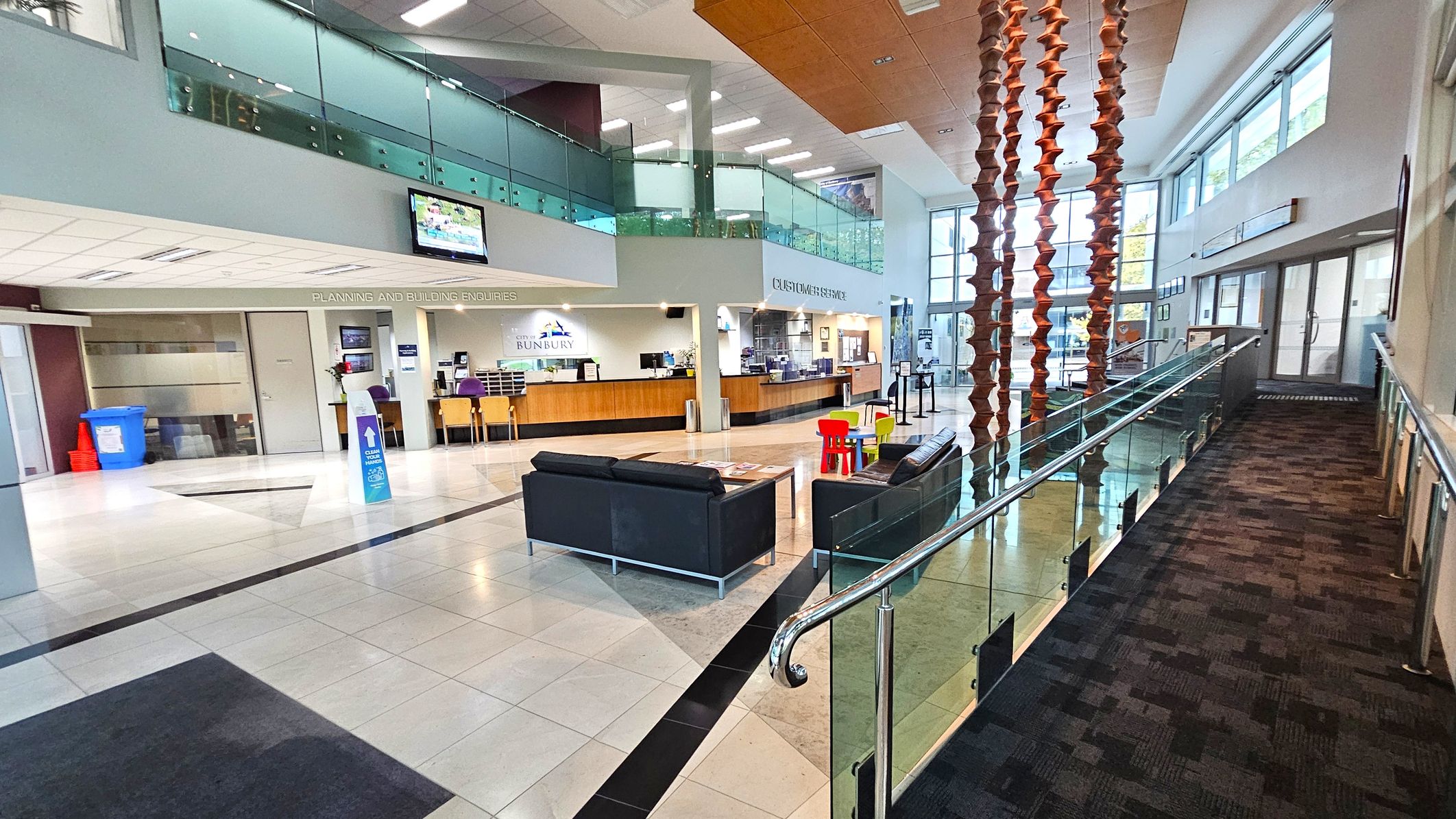 A spacious interior with a modern design. On the left, there is a reception area with a counter and chairs, labelled "Planning and Building Enquiries". Above, a glass-panelled balcony overlooks the space. In the centre, black sofas and a small table with colourful children's chairs are arranged on a tiled floor. On the right, a carpeted ramp with glass railings leads to a set of glass doors. Large windows on the right wall allow natural light to fill the area. A spacious interior with a modern design. On the left, there is a reception area with a counter and chairs, labelled "Planning and Building Enquiries". Above, a glass-panelled balcony overlooks the space. In the centre, black sofas and a small table with colourful children's chairs are arranged on a tiled floor. On the right, a carpeted ramp with glass railings leads to a set of glass doors. Large windows on the right wall allow natural light to fill the area.