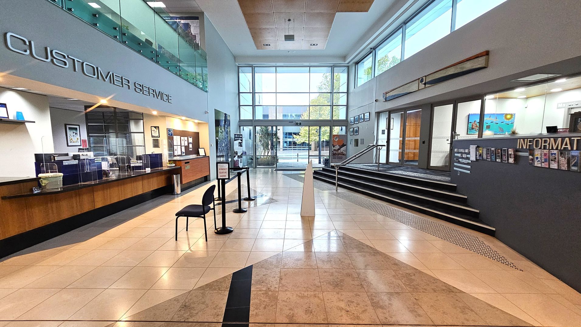 A spacious lobby with a tiled floor. On the left, a customer service area with a long wooden counter and protective screens. Above, a sign reads "CUSTOMER SERVICE". Two chairs are placed near a queue stand. On the right, an information desk with brochures and a sign. A staircase leads up to a glass entrance with large windows, allowing natural light to fill the space. The ceiling is high with recessed lighting. A spacious lobby with a tiled floor. On the left, a customer service area with a long wooden counter and protective screens. Above, a sign reads "CUSTOMER SERVICE". Two chairs are placed near a queue stand. On the right, an information desk with brochures and a sign. A staircase leads up to a glass entrance with large windows, allowing natural light to fill the space. The ceiling is high with recessed lighting.