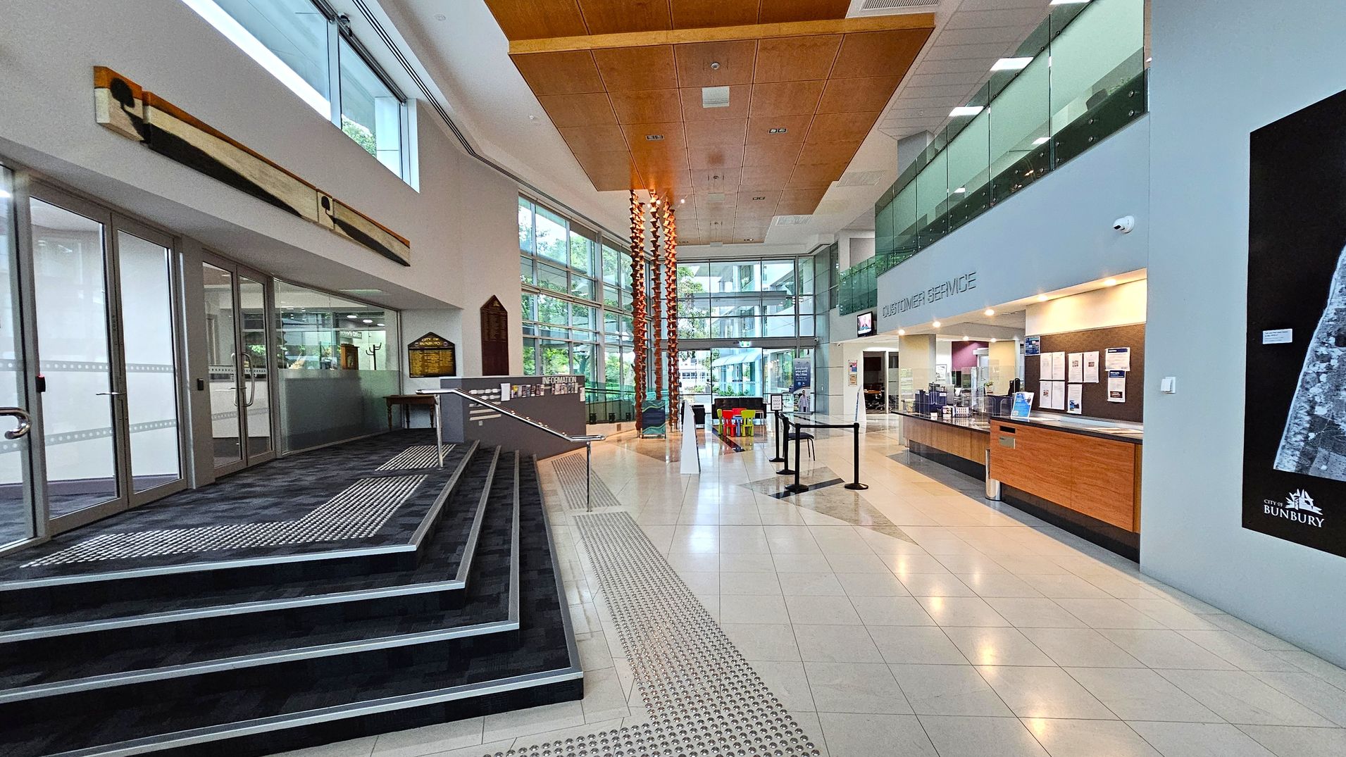 A spacious interior with a modern design. On the left, there are glass doors and a ramp with steps leading up. A long, abstract artwork is mounted on the wall above. In the centre, a tall, decorative column reaches up to the wooden ceiling. On the right, a customer service desk with a wooden counter is visible, along with a large map on the wall. The floor is tiled, and large windows in the background allow natural light to fill the space. A spacious interior with a modern design. On the left, there are glass doors and a ramp with steps leading up. A long, abstract artwork is mounted on the wall above. In the centre, a tall, decorative column reaches up to the wooden ceiling. On the right, a customer service desk with a wooden counter is visible, along with a large map on the wall. The floor is tiled, and large windows in the background allow natural light to fill the space.