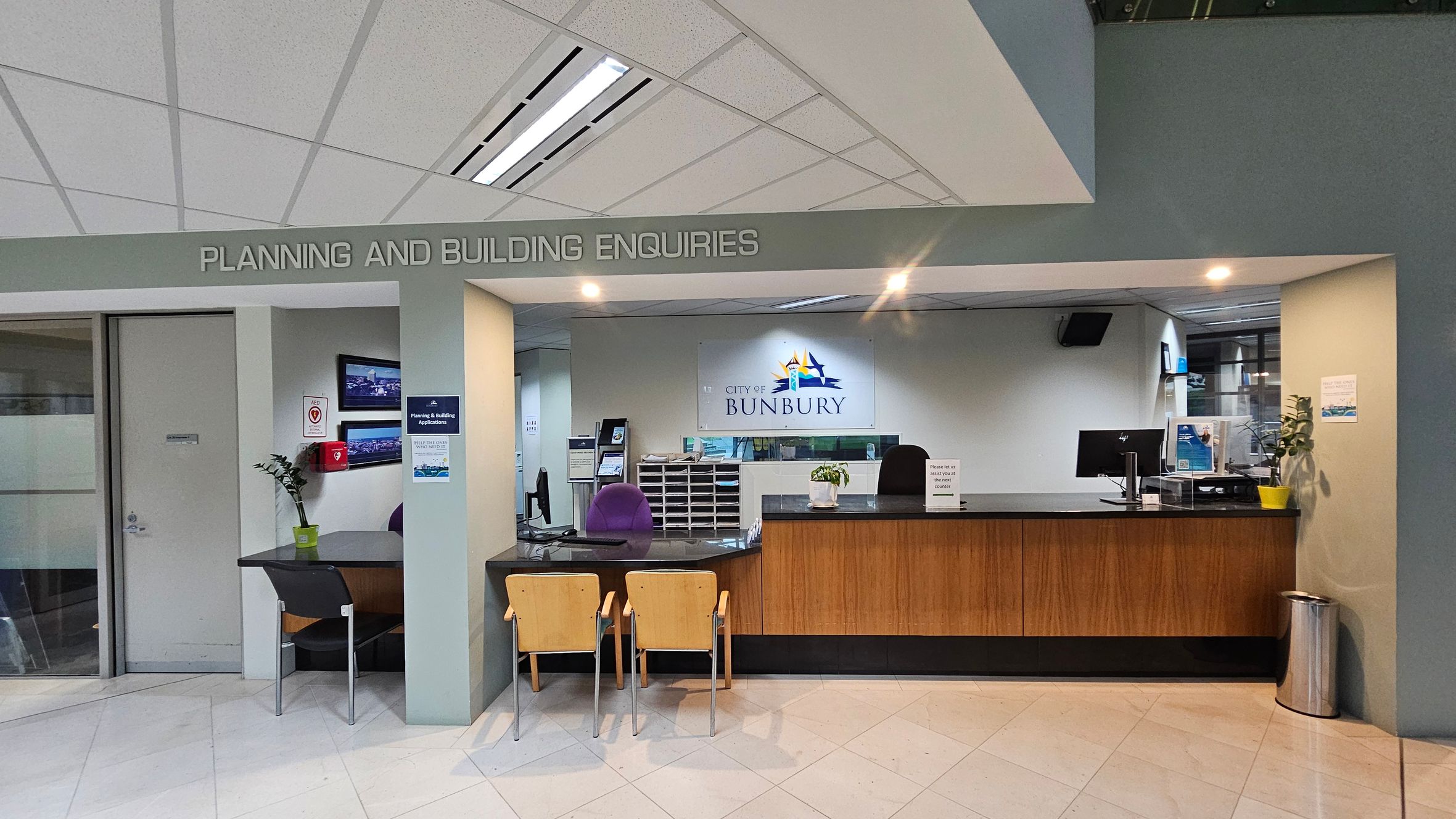 A reception area with a sign above reading "PLANNING AND BUILDING ENQUIRIES". On the left, there's a small table with a chair, a plant, and a wall-mounted screen. A door is adjacent to this setup. In the centre, a counter with two wooden chairs faces a desk with a computer and office supplies. Behind the counter, a sign reads "CITY OF BUNBURY". The floor is tiled, and the ceiling has a grid pattern with fluorescent lights. A small plant and a bin are on the right side of the counter. A reception area with a sign above reading "PLANNING AND BUILDING ENQUIRIES". On the left, there's a small table with a chair, a plant, and a wall-mounted screen. A door is adjacent to this setup. In the centre, a counter with two wooden chairs faces a desk with a computer and office supplies. Behind the counter, a sign reads "CITY OF BUNBURY". The floor is tiled, and the ceiling has a grid pattern with fluorescent lights. A small plant and a bin are on the right side of the counter.