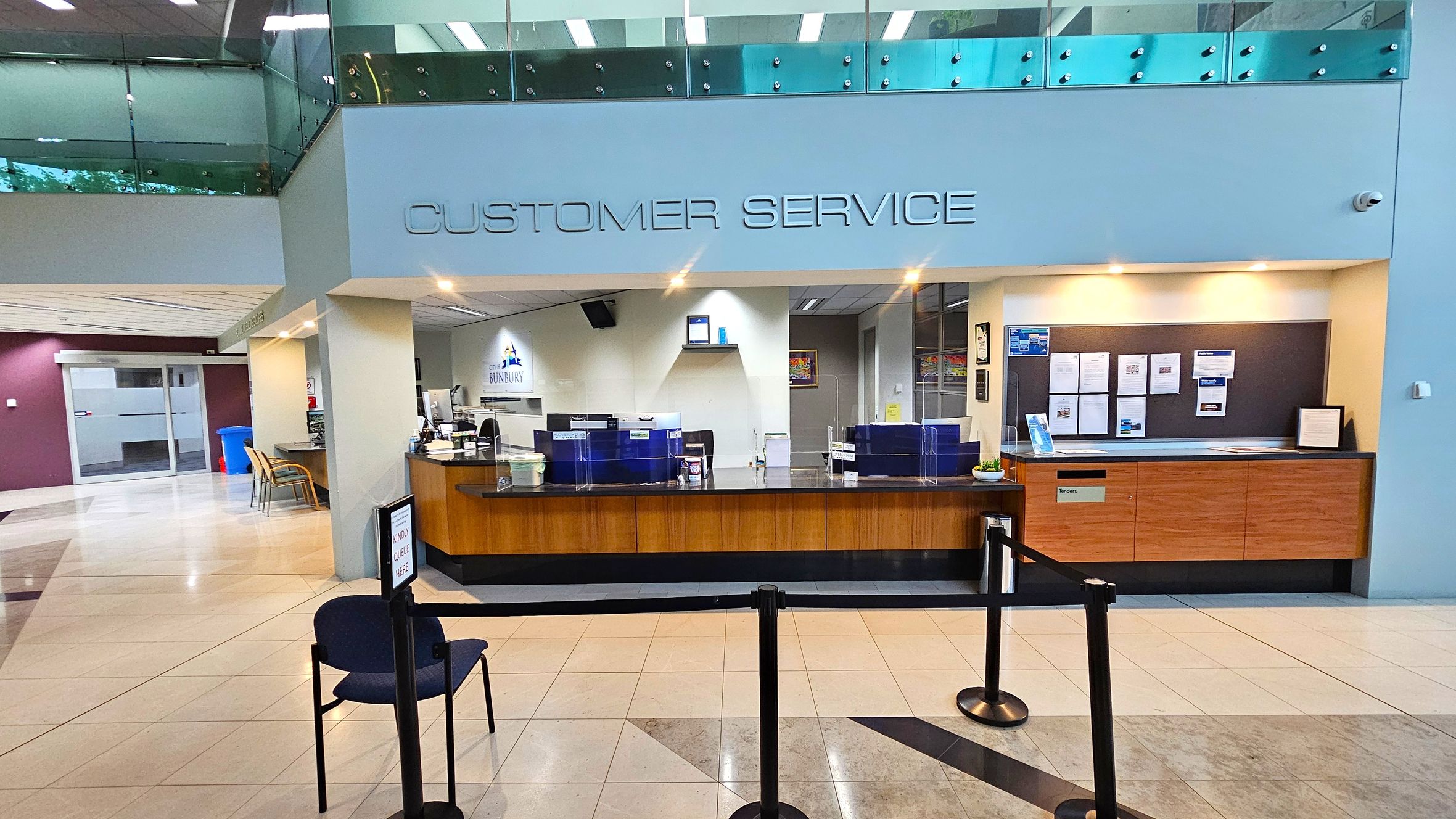 A customer service area with a wooden counter and clear partitions. Above, "CUSTOMER SERVICE" is written on the wall. Behind the counter, there are computers and a sign with "BUNBURY" visible. On the right, a notice board displays various papers. The floor is tiled, and a single chair is placed in front of a queue barrier. The ceiling has recessed lighting, and a glass railing is visible on the upper level. A customer service area with a wooden counter and clear partitions. Above, "CUSTOMER SERVICE" is written on the wall. Behind the counter, there are computers and a sign with "BUNBURY" visible. On the right, a notice board displays various papers. The floor is tiled, and a single chair is placed in front of a queue barrier. The ceiling has recessed lighting, and a glass railing is visible on the upper level.