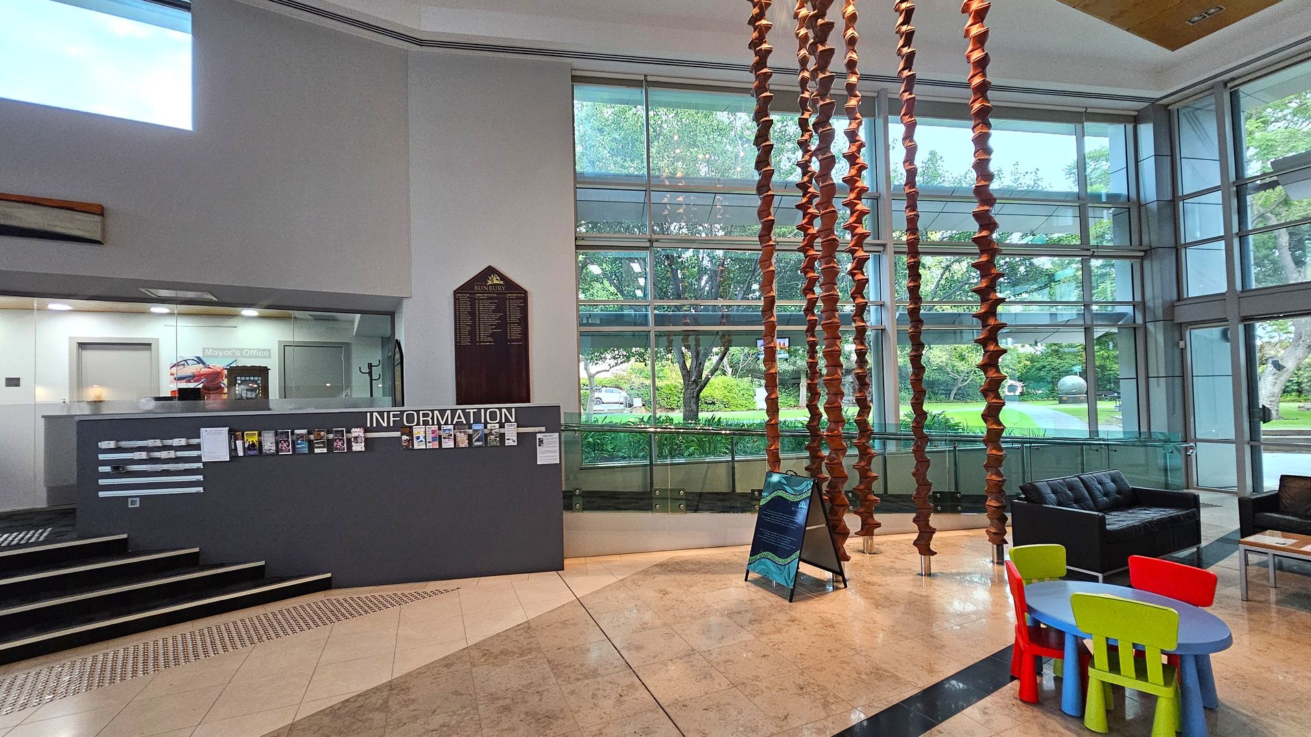 A spacious interior with a tiled floor and high ceiling. On the left, there's an information desk with brochures and a sign for the Mayor's Office. Behind the desk, a glass wall and door are visible. In the centre, a decorative hanging installation made of stacked, brown elements. On the right, a seating area with a black sofa and a small children's table with colourful chairs. Large windows in the background offer a view of greenery outside. A spacious interior with a tiled floor and high ceiling. On the left, there's an information desk with brochures and a sign for the Mayor's Office. Behind the desk, a glass wall and door are visible. In the centre, a decorative hanging installation made of stacked, brown elements. On the right, a seating area with a black sofa and a small children's table with colourful chairs. Large windows in the background offer a view of greenery outside.