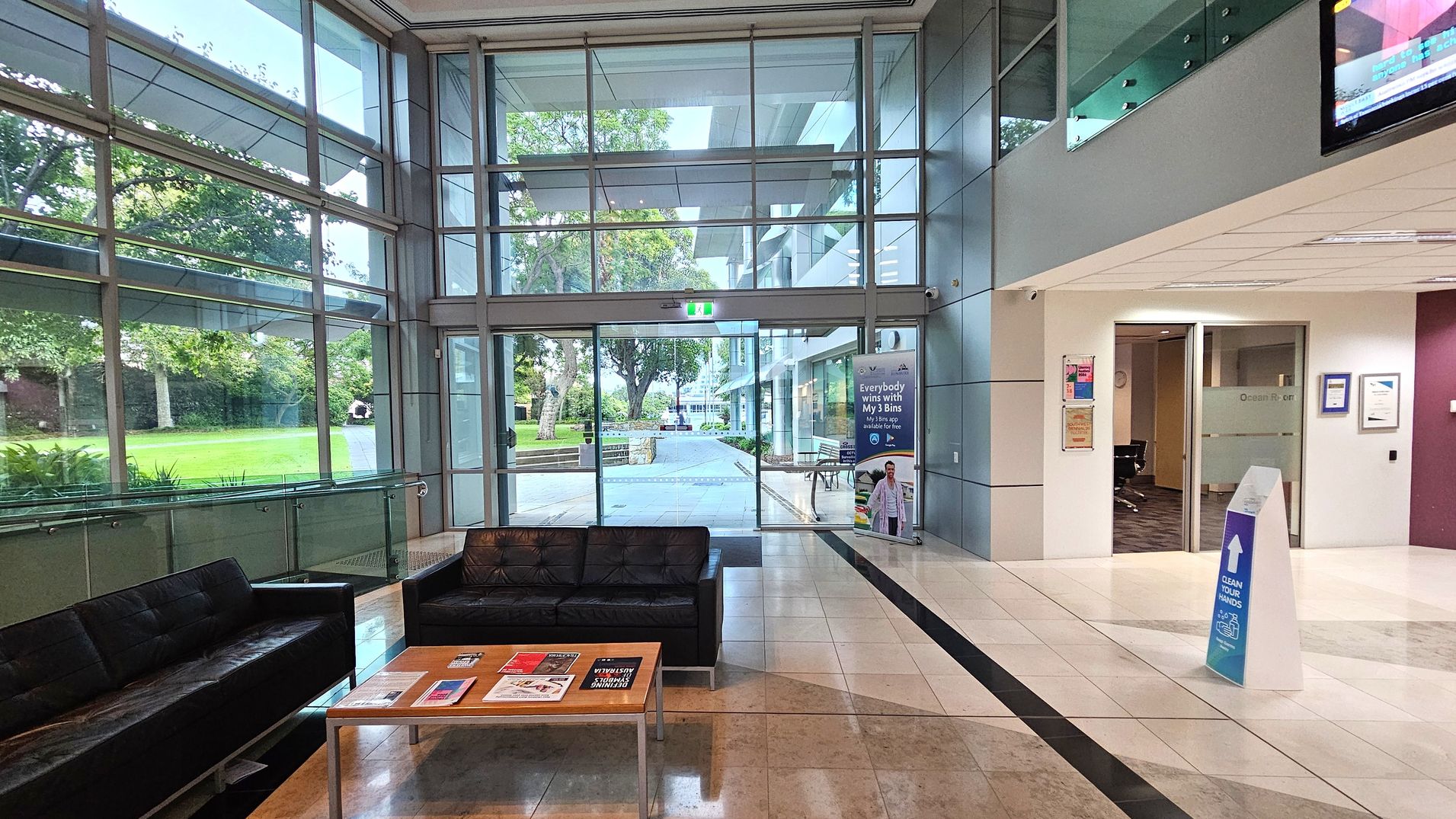 A modern lobby area with large glass windows on the left, offering a view of a green lawn and trees outside. In the centre, there is a glass entrance door with a safety exit sign above. To the right, a white wall with an open doorway leading to an office space. A black leather sofa and a wooden coffee table with magazines are in the foreground. The floor is tiled in a light colour with a dark strip, and a sign on the floor advises to clean hands. A modern lobby area with large glass windows on the left, offering a view of a green lawn and trees outside. In the centre, there is a glass entrance door with a safety exit sign above. To the right, a white wall with an open doorway leading to an office space. A black leather sofa and a wooden coffee table with magazines are in the foreground. The floor is tiled in a light colour with a dark strip, and a sign on the floor advises to clean hands.
