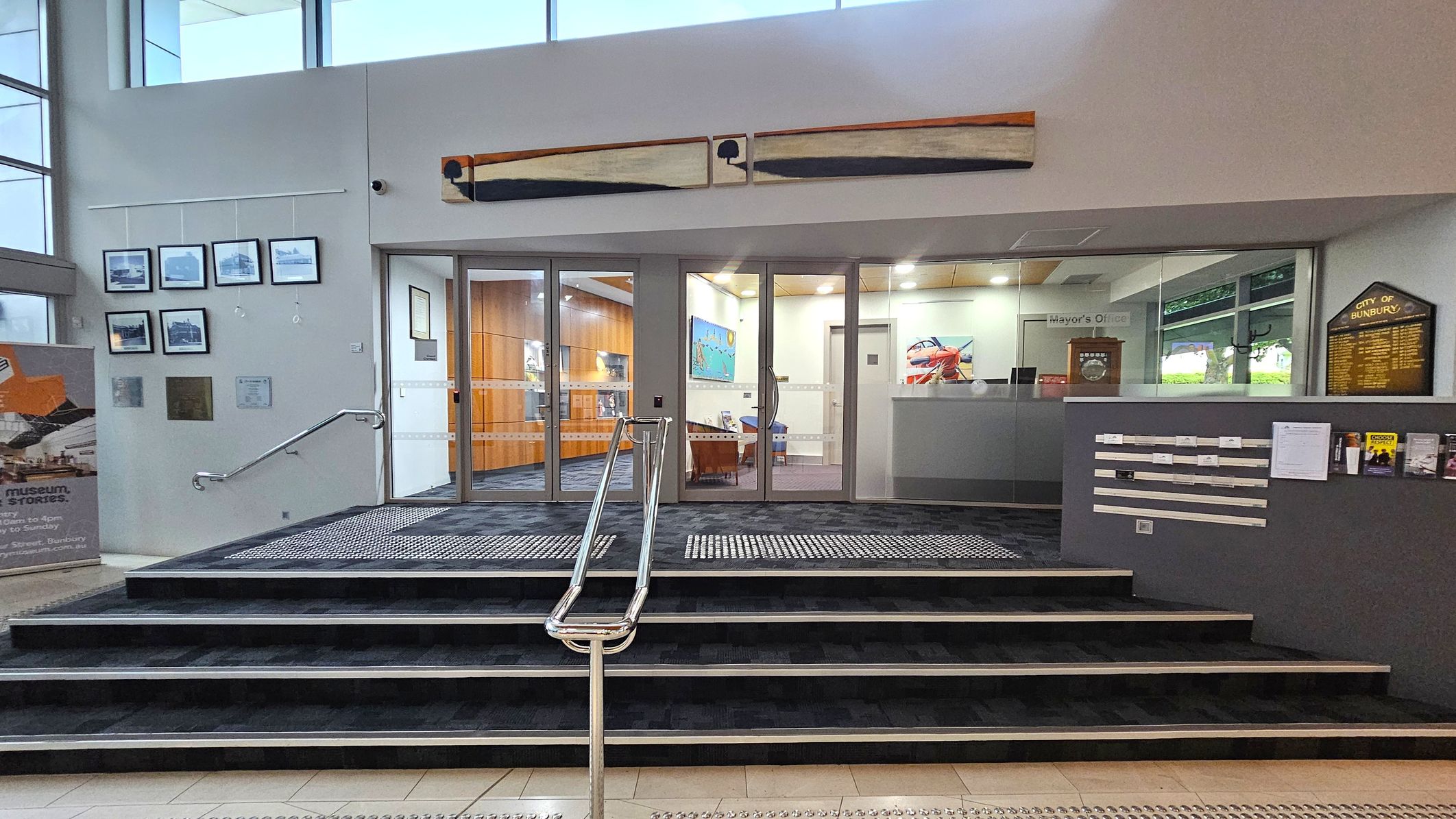 A modern interior with a set of stairs leading up to a glass entrance. On the left, framed photographs are displayed on the wall above a handrail. A sign with text is positioned on the floor. Above the entrance, a long artwork is mounted. To the right, a grey wall features a plaque and several brochures. Inside, the space is well-lit with wooden panelling and colourful artwork visible. A sign indicates the "Mayor's Office". The floor is tiled with a textured mat at the entrance. A modern interior with a set of stairs leading up to a glass entrance. On the left, framed photographs are displayed on the wall above a handrail. A sign with text is positioned on the floor. Above the entrance, a long artwork is mounted. To the right, a grey wall features a plaque and several brochures. Inside, the space is well-lit with wooden panelling and colourful artwork visible. A sign indicates the "Mayor's Office". The floor is tiled with a textured mat at the entrance.