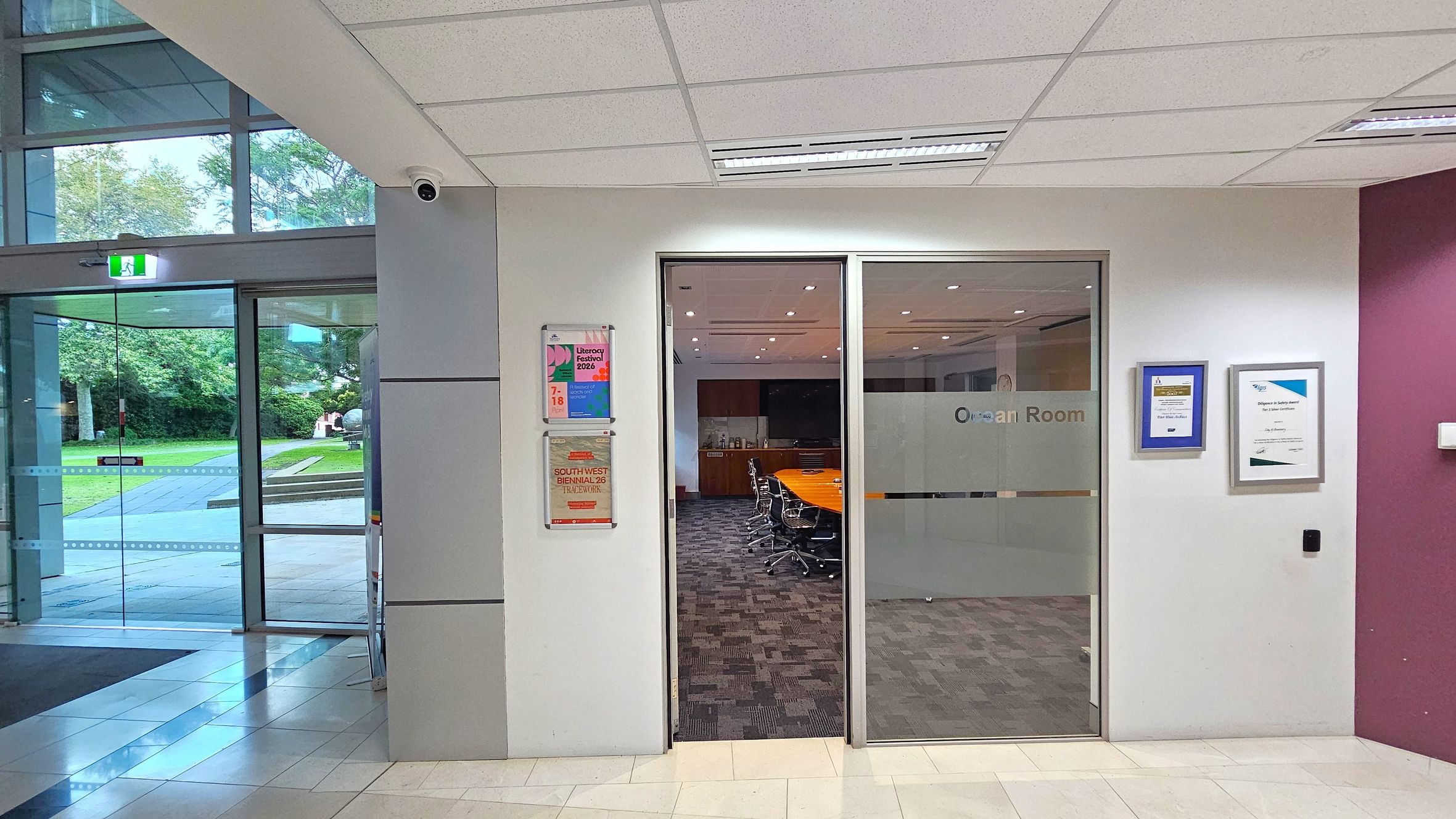 A hallway with a glass door on the left leading outside, showing a grassy area and trees. To the right, there is a glass entrance to a room labelled "Ocean Room," with a conference table and chairs visible inside. The wall beside the door has posters and certificates displayed. The floor is tiled, and the ceiling has recessed lighting and a security camera. A hallway with a glass door on the left leading outside, showing a grassy area and trees. To the right, there is a glass entrance to a room labelled "Ocean Room," with a conference table and chairs visible inside. The wall beside the door has posters and certificates displayed. The floor is tiled, and the ceiling has recessed lighting and a security camera.