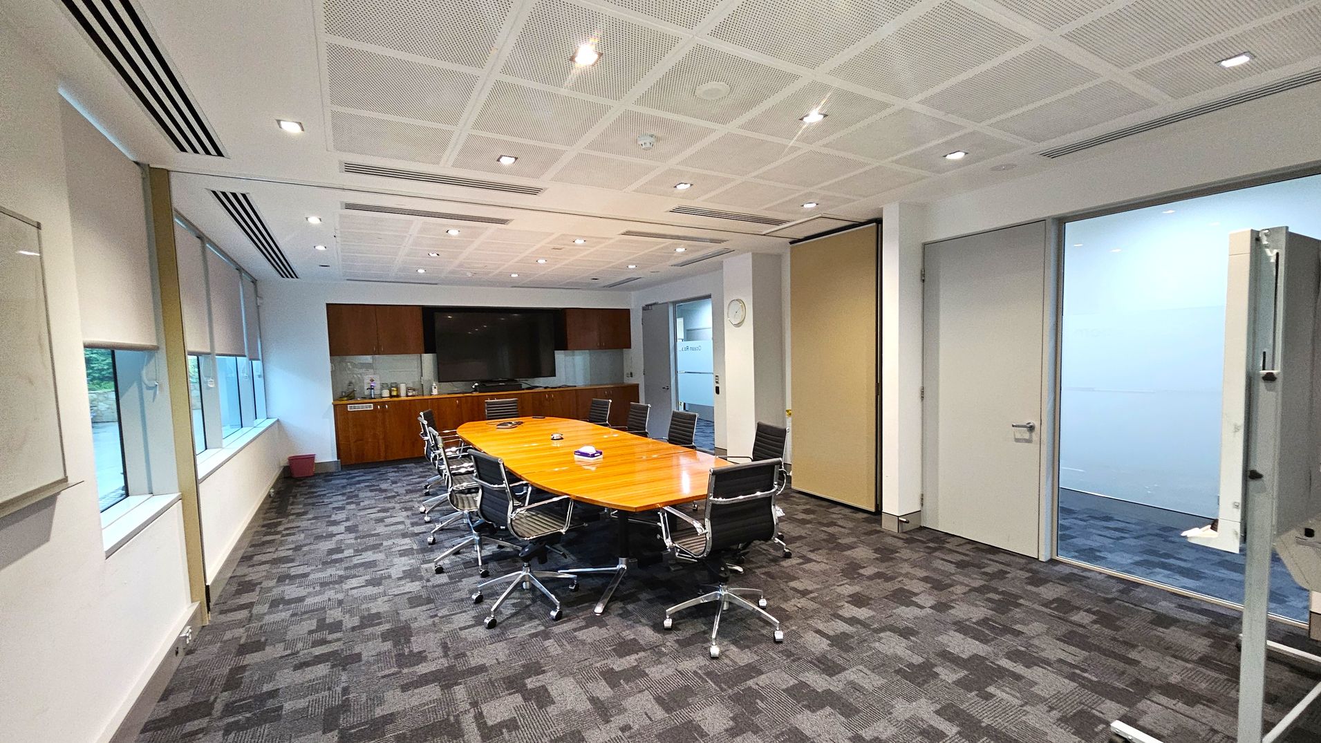 A modern conference room with a long wooden table in the centre, surrounded by black office chairs on wheels. On the left, large windows with blinds allow natural light in. The back wall features a large screen and a wooden cabinet with various items. The right side has a door leading to another room and a whiteboard. The floor is carpeted with a grey and purple pattern, and the ceiling is white with recessed lighting. A modern conference room with a long wooden table in the centre, surrounded by black office chairs on wheels. On the left, large windows with blinds allow natural light in. The back wall features a large screen and a wooden cabinet with various items. The right side has a door leading to another room and a whiteboard. The floor is carpeted with a grey and purple pattern, and the ceiling is white with recessed lighting.
