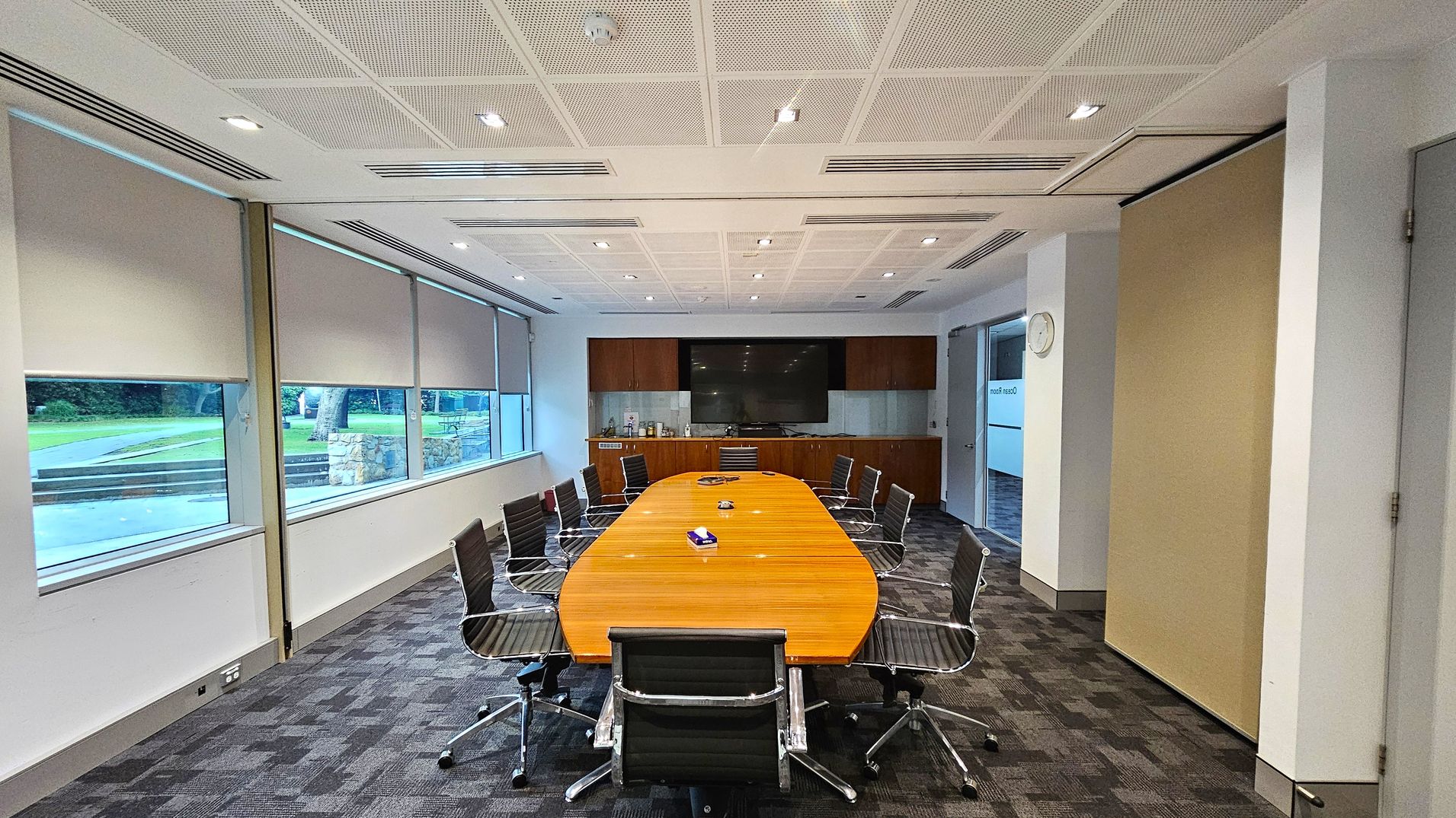 A conference room with a long, oval wooden table surrounded by black office chairs on a patterned carpet floor. The left side features large windows with roller blinds partially down, revealing a view of a grassy outdoor area. The back wall has a large flat-screen TV mounted above a wooden cabinet. The ceiling is white with recessed lighting. On the right, a wall clock is visible above a doorway. A conference room with a long, oval wooden table surrounded by black office chairs on a patterned carpet floor. The left side features large windows with roller blinds partially down, revealing a view of a grassy outdoor area. The back wall has a large flat-screen TV mounted above a wooden cabinet. The ceiling is white with recessed lighting. On the right, a wall clock is visible above a doorway.