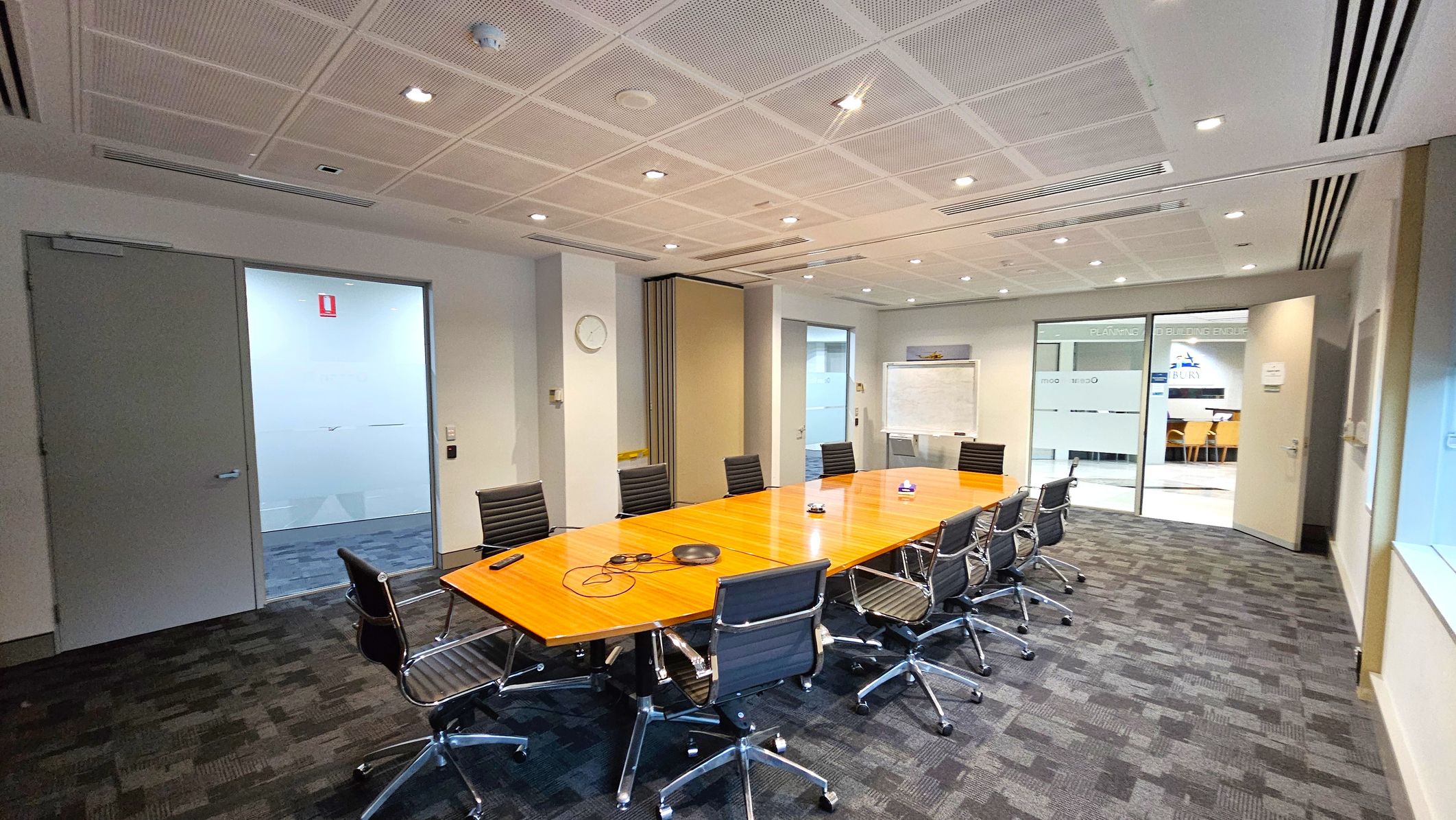 A conference room with a long wooden table surrounded by black office chairs on a dark carpeted floor. On the left, there is a closed door and a wall clock. In the centre, a whiteboard is visible against the wall. On the right, glass doors lead to another area with visible signage and a glimpse of a seating area. The ceiling is fitted with recessed lighting and air vents. A conference room with a long wooden table surrounded by black office chairs on a dark carpeted floor. On the left, there is a closed door and a wall clock. In the centre, a whiteboard is visible against the wall. On the right, glass doors lead to another area with visible signage and a glimpse of a seating area. The ceiling is fitted with recessed lighting and air vents.