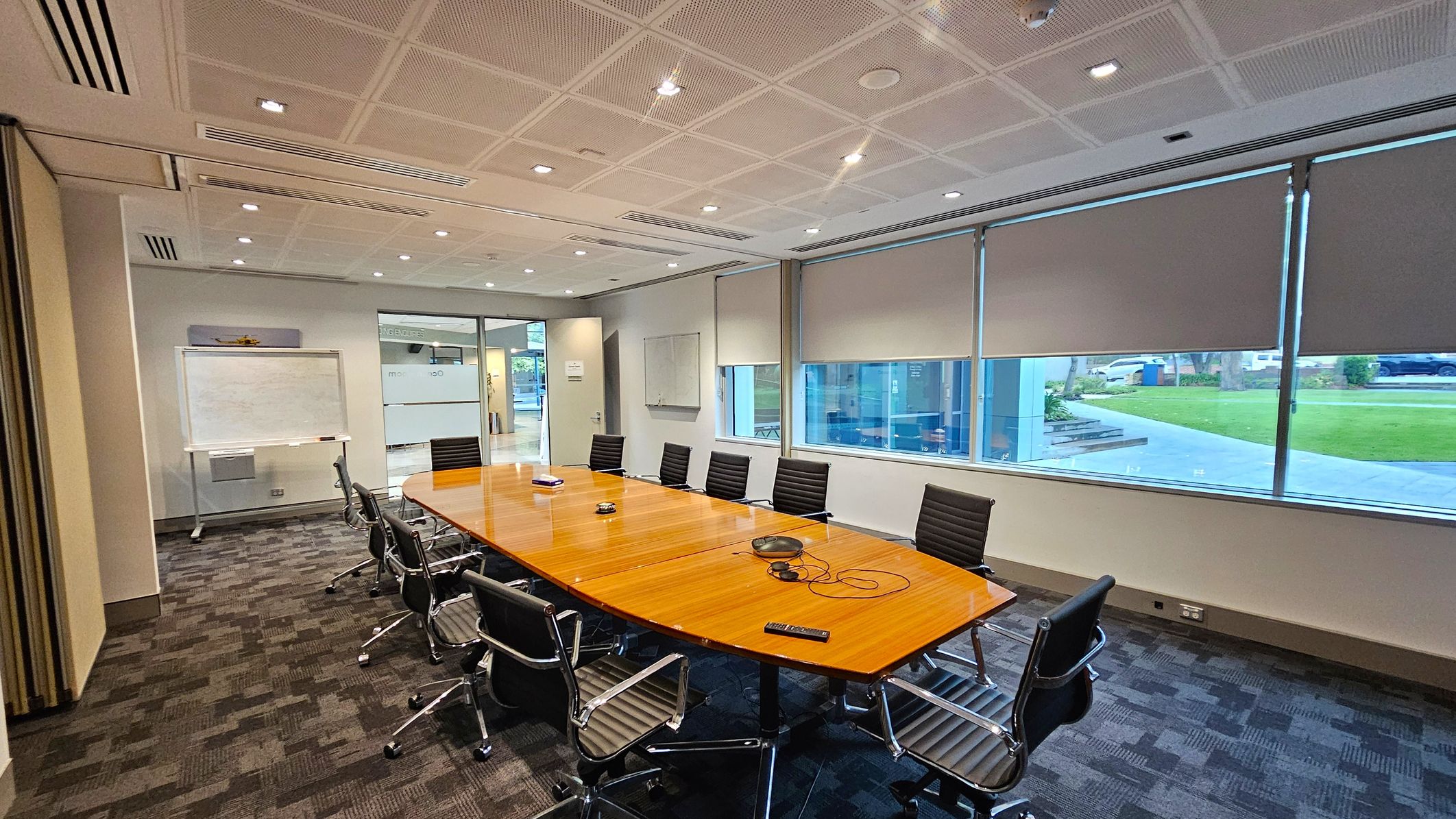 A modern conference room with a large wooden table in the centre surrounded by black office chairs on wheels. On the left, there's a whiteboard on a stand. The right side features large windows with partially drawn blinds, offering a view of a grassy area outside. The floor is carpeted with a grey and black pattern, and the ceiling is fitted with recessed lighting and air vents. A modern conference room with a large wooden table in the centre surrounded by black office chairs on wheels. On the left, there's a whiteboard on a stand. The right side features large windows with partially drawn blinds, offering a view of a grassy area outside. The floor is carpeted with a grey and black pattern, and the ceiling is fitted with recessed lighting and air vents.