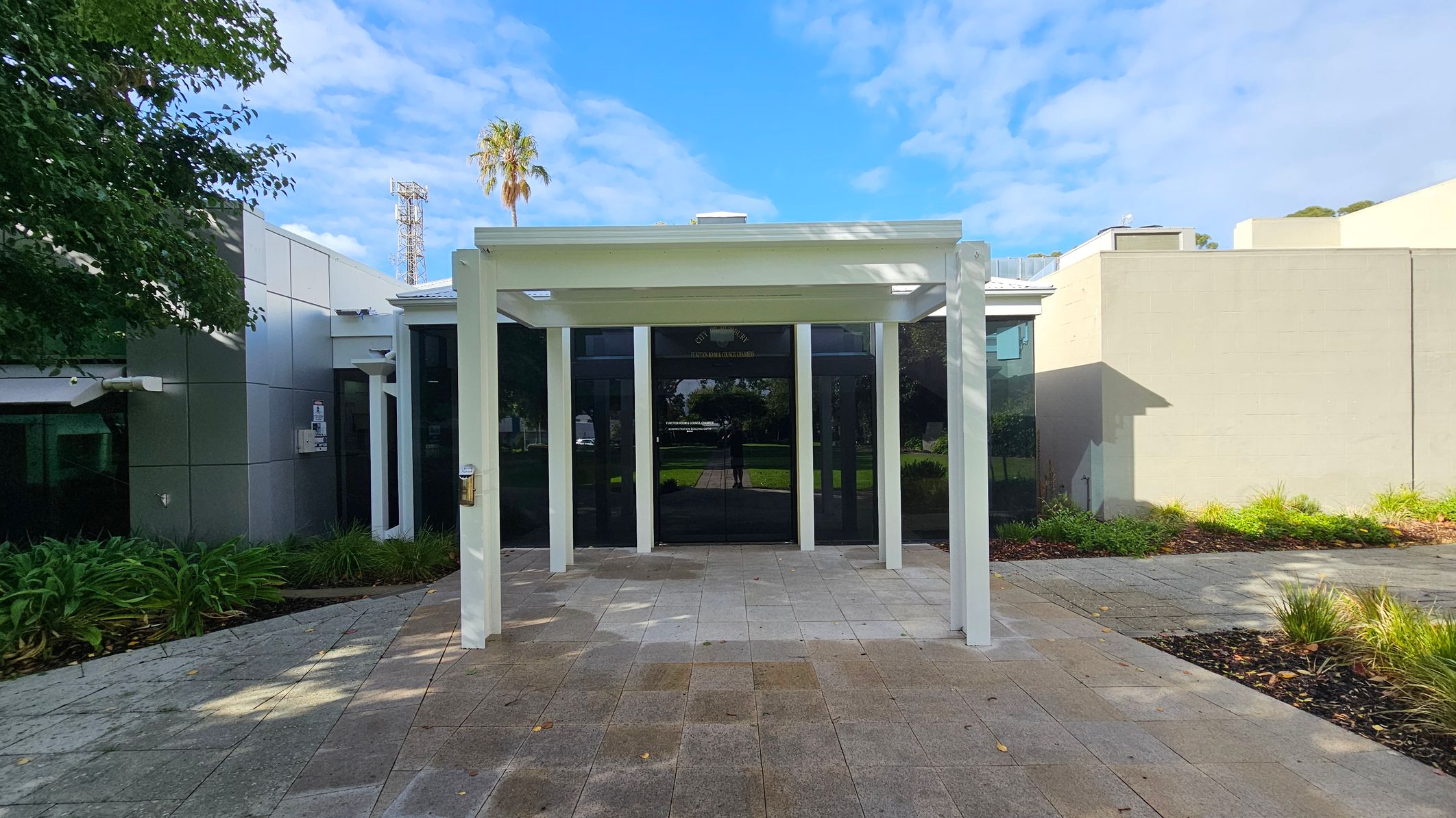 A modern building entrance with a white pergola structure in the foreground. The ground is paved with grey and brown tiles. On the left, there are green plants and a tree partially visible. The building has a glass facade with reflections and a light grey wall on the right. In the background, a palm tree and a tall metal structure are visible against a blue sky. A modern building entrance with a white pergola structure in the foreground. The ground is paved with grey and brown tiles. On the left, there are green plants and a tree partially visible. The building has a glass facade with reflections and a light grey wall on the right. In the background, a palm tree and a tall metal structure are visible against a blue sky.