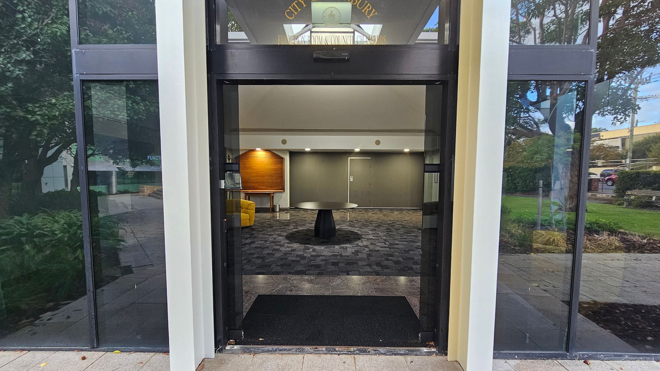 A glass entrance with a black mat on the floor leads into a building. Inside, there's a round black table on a patterned carpet. To the left, a yellow chair and a wooden board are visible. The walls are grey with a door in the centre. Outside, reflections of trees and greenery are seen on the glass panels. The ceiling inside is plain with recessed lighting. A glass entrance with a black mat on the floor leads into a building. Inside, there's a round black table on a patterned carpet. To the left, a yellow chair and a wooden board are visible. The walls are grey with a door in the centre. Outside, reflections of trees and greenery are seen on the glass panels. The ceiling inside is plain with recessed lighting.