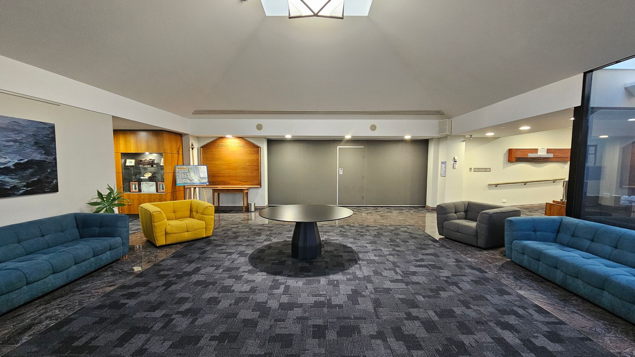 A spacious room with a patterned dark carpet. In the centre, a round black table stands on the floor. On the left, a blue sofa and a yellow armchair are placed near a wooden display case and a painting. On the right, another blue sofa and a grey armchair are positioned near a wall with a wooden shelf. The back wall features a wooden panel and a closed door. The ceiling is light-coloured with a skylight, and the room is well-lit with recessed lighting. A spacious room with a patterned dark carpet. In the centre, a round black table stands on the floor. On the left, a blue sofa and a yellow armchair are placed near a wooden display case and a painting. On the right, another blue sofa and a grey armchair are positioned near a wall with a wooden shelf. The back wall features a wooden panel and a closed door. The ceiling is light-coloured with a skylight, and the room is well-lit with recessed lighting.