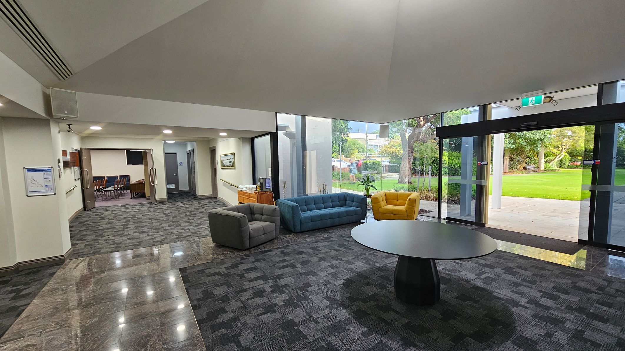 A spacious lobby area with a grey carpeted floor. On the left, there is an open doorway leading to a room with chairs. A grey armchair, a blue sofa, and a yellow armchair are arranged around a circular black table in the centre. Large glass doors on the right open to a green outdoor area with trees. The ceiling is high and light-coloured, with recessed lighting. A wooden cabinet with a coffee machine is against the wall near the glass doors. A spacious lobby area with a grey carpeted floor. On the left, there is an open doorway leading to a room with chairs. A grey armchair, a blue sofa, and a yellow armchair are arranged around a circular black table in the centre. Large glass doors on the right open to a green outdoor area with trees. The ceiling is high and light-coloured, with recessed lighting. A wooden cabinet with a coffee machine is against the wall near the glass doors.