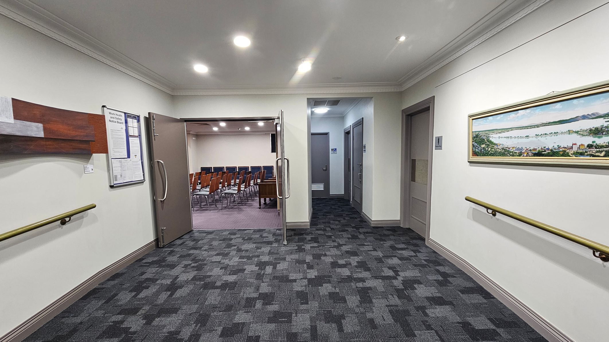 A hallway with a patterned carpet floor and white walls. On the left, there's a wooden notice board and a handrail. A door is open, leading to a room with rows of wooden chairs. On the right, a framed panoramic picture hangs above another handrail. Two closed doors are visible further down the hallway. The ceiling has recessed lighting. A hallway with a patterned carpet floor and white walls. On the left, there's a wooden notice board and a handrail. A door is open, leading to a room with rows of wooden chairs. On the right, a framed panoramic picture hangs above another handrail. Two closed doors are visible further down the hallway. The ceiling has recessed lighting.