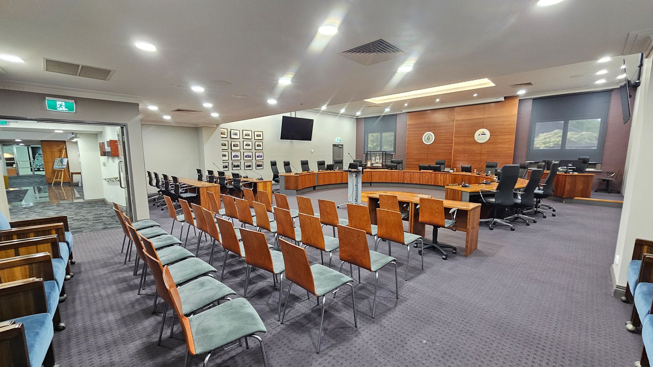 A spacious conference room with rows of wooden chairs with green cushions arranged in the foreground. To the right, a large U-shaped wooden table is surrounded by black swivel chairs. The back wall features wood panelling with two emblems and a large screen. On the left, there are framed pictures on the wall and an open doorway leading to another area. The ceiling is fitted with recessed lighting, and the floor is carpeted in a dark grey pattern. A spacious conference room with rows of wooden chairs with green cushions arranged in the foreground. To the right, a large U-shaped wooden table is surrounded by black swivel chairs. The back wall features wood panelling with two emblems and a large screen. On the left, there are framed pictures on the wall and an open doorway leading to another area. The ceiling is fitted with recessed lighting, and the floor is carpeted in a dark grey pattern.