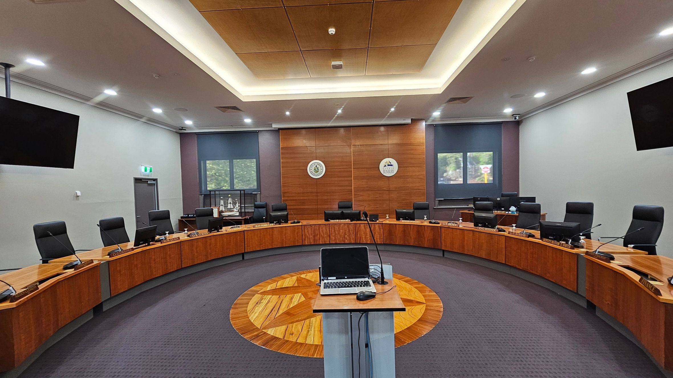 A modern conference room with a semi-circular arrangement of wooden desks and black chairs. In the centre, a podium with a laptop and microphone stands on a patterned wooden floor. The back wall features two emblems and two windows with blue blinds. The ceiling has recessed lighting and a central wooden panel. Two large screens are mounted on the side walls. The floor is carpeted in grey. A modern conference room with a semi-circular arrangement of wooden desks and black chairs. In the centre, a podium with a laptop and microphone stands on a patterned wooden floor. The back wall features two emblems and two windows with blue blinds. The ceiling has recessed lighting and a central wooden panel. Two large screens are mounted on the side walls. The floor is carpeted in grey.