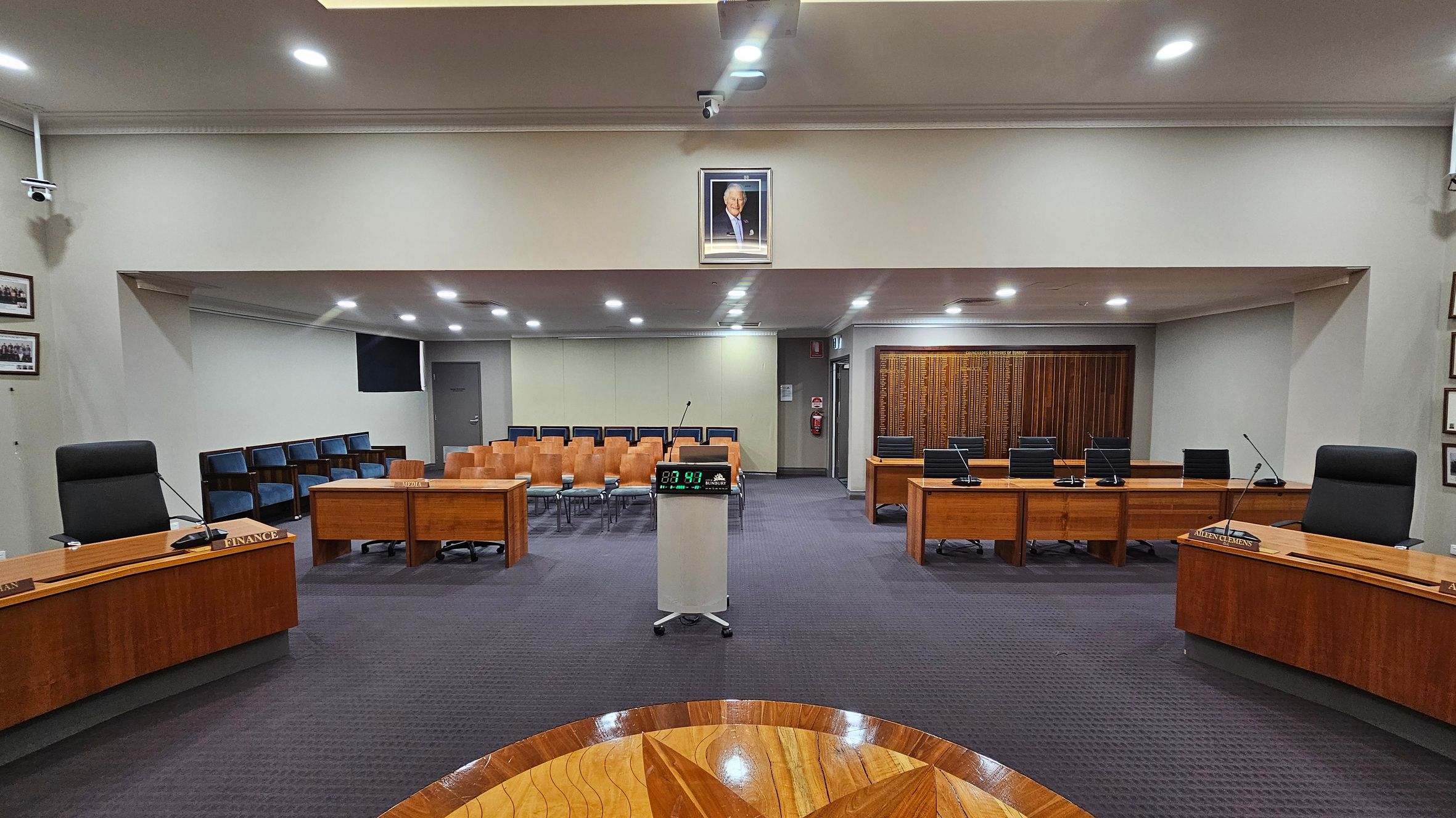 A council chamber with a central podium featuring a digital timer. Surrounding the podium are wooden desks with microphones and nameplates. On the left and right are black chairs behind the desks. In the background, rows of wooden and blue chairs face the front. A framed portrait hangs above the central area. The floor is carpeted in dark grey, and the ceiling has recessed lighting. Walls are adorned with framed photos and a wooden panel. A council chamber with a central podium featuring a digital timer. Surrounding the podium are wooden desks with microphones and nameplates. On the left and right are black chairs behind the desks. In the background, rows of wooden and blue chairs face the front. A framed portrait hangs above the central area. The floor is carpeted in dark grey, and the ceiling has recessed lighting. Walls are adorned with framed photos and a wooden panel.