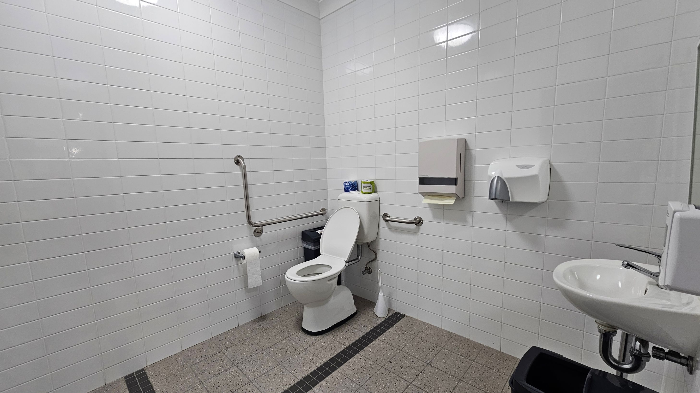 A public restroom with white tiled walls and a grey tiled floor. On the left, there is a toilet with a raised seat, equipped with grab bars on both sides. Above the toilet, there are two rolls of toilet paper. A toilet brush is on the floor beside it. On the right wall, there is a paper towel dispenser and a soap dispenser above a small sink. Below the sink, a black waste bin is visible. The ceiling has a simple light fixture. A public restroom with white tiled walls and a grey tiled floor. On the left, there is a toilet with a raised seat, equipped with grab bars on both sides. Above the toilet, there are two rolls of toilet paper. A toilet brush is on the floor beside it. On the right wall, there is a paper towel dispenser and a soap dispenser above a small sink. Below the sink, a black waste bin is visible. The ceiling has a simple light fixture.