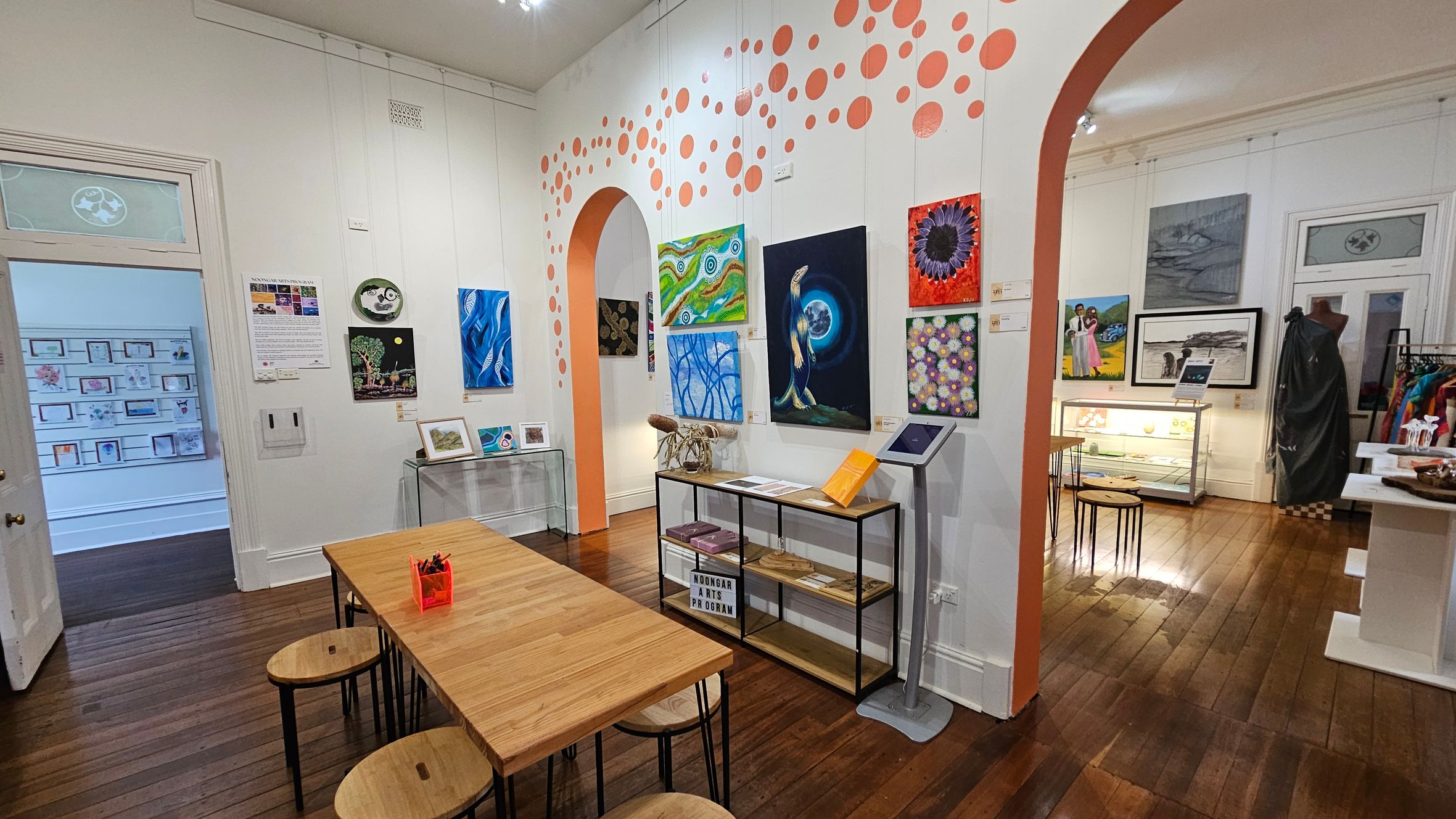 A gallery room with wooden flooring and white walls adorned with various artworks. On the left, a doorway leads to another room displaying framed pictures. A long wooden table with stools is in the foreground. The centre wall features colourful paintings and a shelf with books and art materials. An archway with orange dots adds a decorative touch. On the right, more artworks are displayed, along with a mannequin and a glass display case. Ceiling lights illuminate the space.