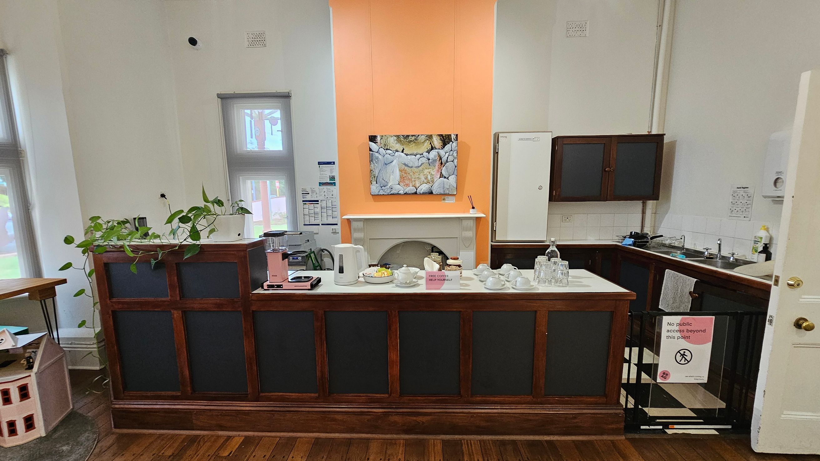 A small kitchen area with a wooden counter in the foreground, topped with a kettle, coffee maker, cups, and a sign offering free coffee. A plant is placed on the left side of the counter. Behind the counter, there's a fireplace with an orange wall above it, displaying a painting. To the right, a sink and cabinets are visible. A sign on a gate reads "No public access beyond this point." The floor is wooden, and the ceiling is not visible.