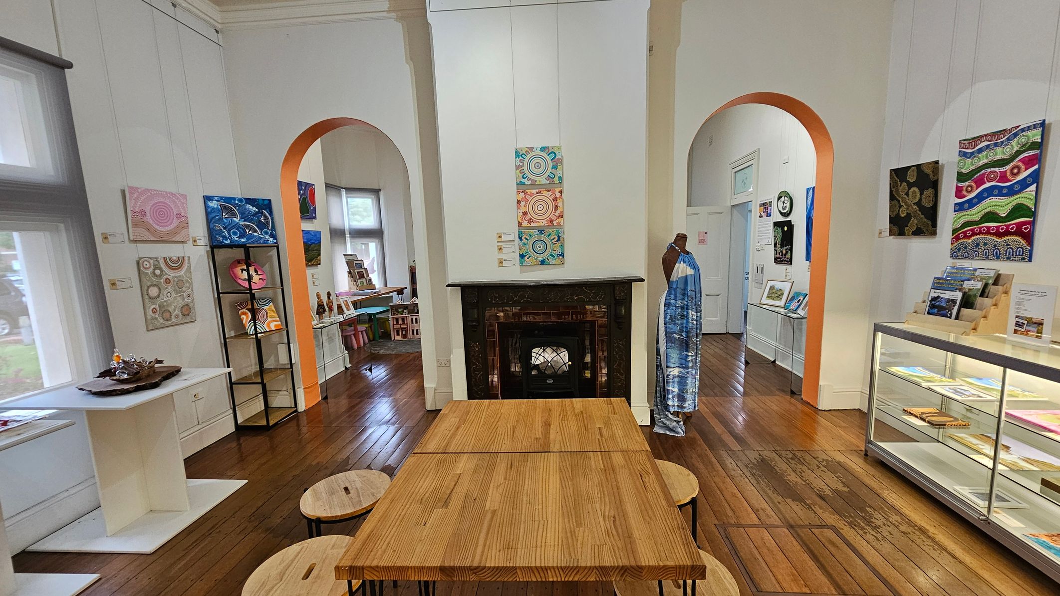 A room with wooden flooring and white walls features a central wooden table with round stools. On the left, a white table displays decorative items, and colourful artwork hangs on the walls. Two arched doorways lead to adjacent rooms. A fireplace with a black mantle is in the centre, with artwork above it. To the right, a mannequin draped in fabric stands near a glass display case filled with books and pamphlets. Bright natural light filters through large windows.