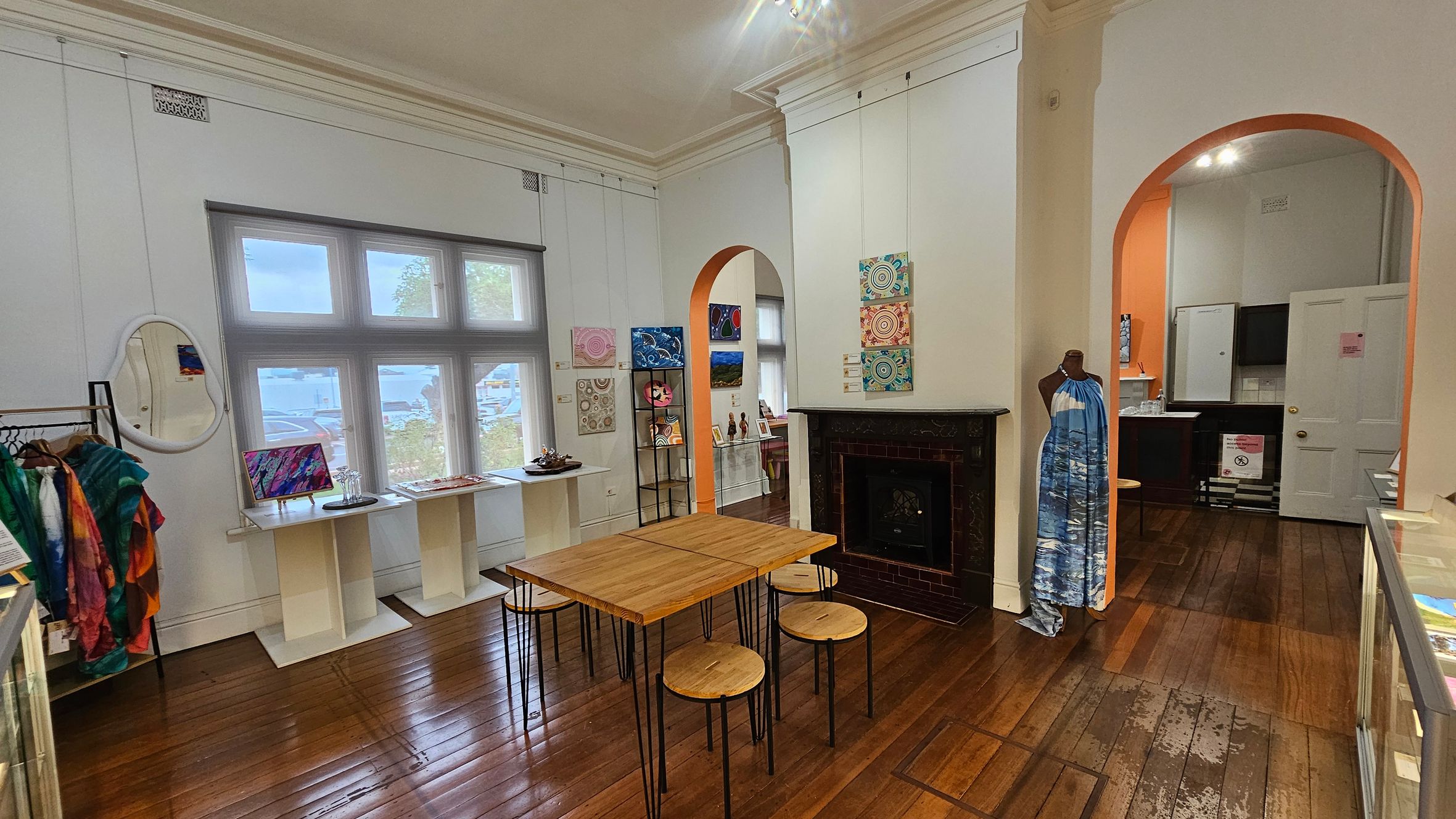 A room with wooden flooring and a high ceiling. On the left, a rack with colourful garments and a mirror. A window with a view of the outside is in the centre, flanked by small tables displaying art and items. A wooden table with stools is in the middle. On the right, a mannequin wearing a blue dress stands near a fireplace. Two archways lead to another room with orange walls and a door. Various artworks are displayed on the walls.