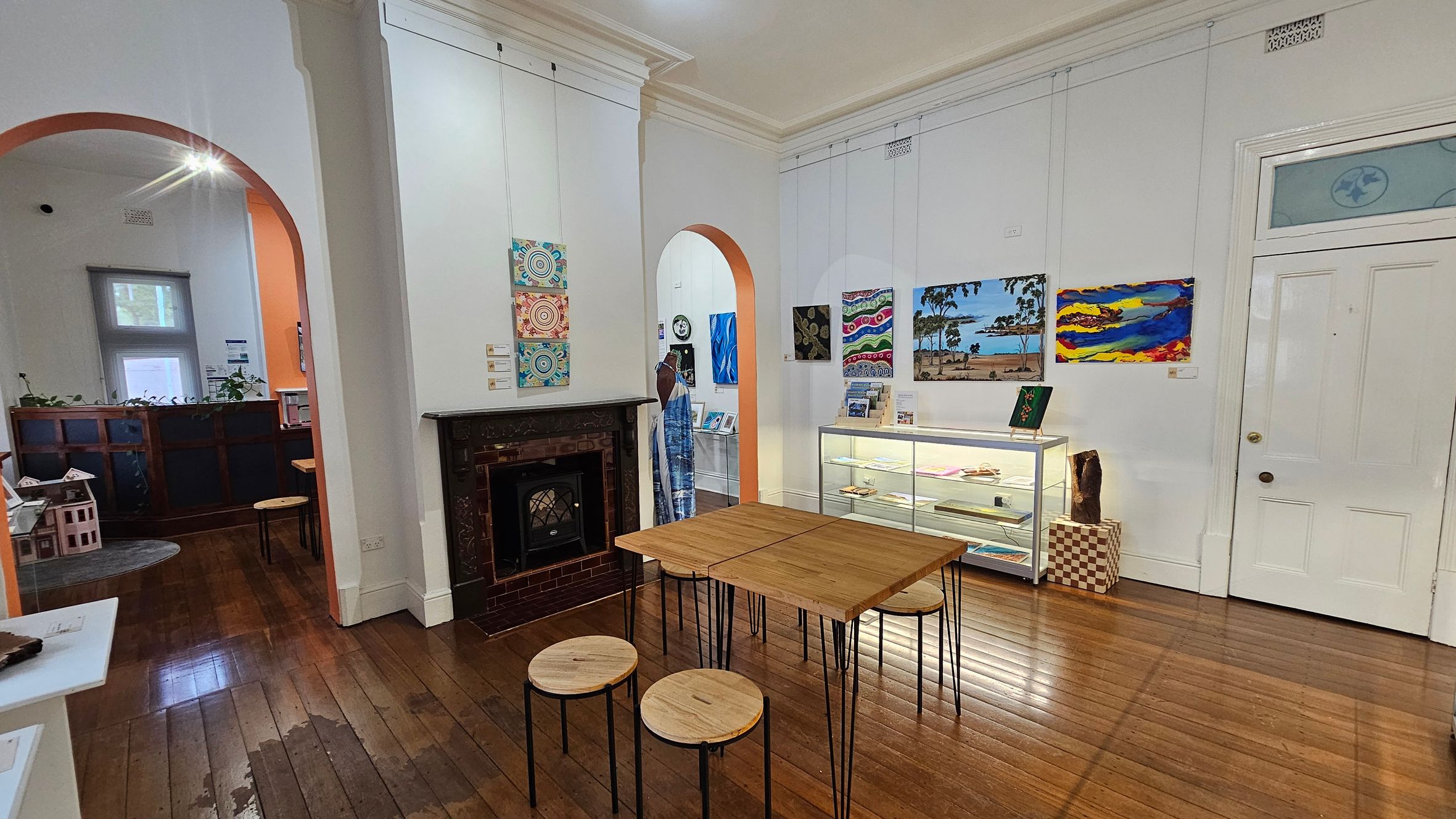 A room with wooden flooring and a high ceiling. In the centre, there is a wooden table with round stools. On the right, a glass display case holds various items, and colourful paintings hang on the wall above it. A white door is visible next to the display. On the left, a fireplace with a dark mantel is set into the wall, and an archway leads to another room with a window and plants on a cabinet.