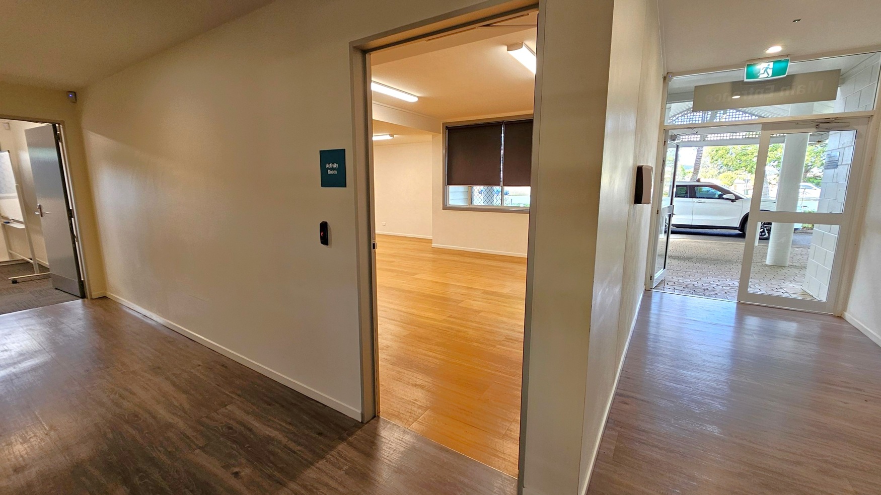 A hallway with dark wood flooring leads to an open door on the left, revealing a room with light wood flooring and a window with a blind. A sign next to the door reads "Activity Room." To the right, a glass door opens to the outside, where a white car is visible on a brick-paved area. The ceiling has fluorescent lights, and an exit sign is above the glass door.