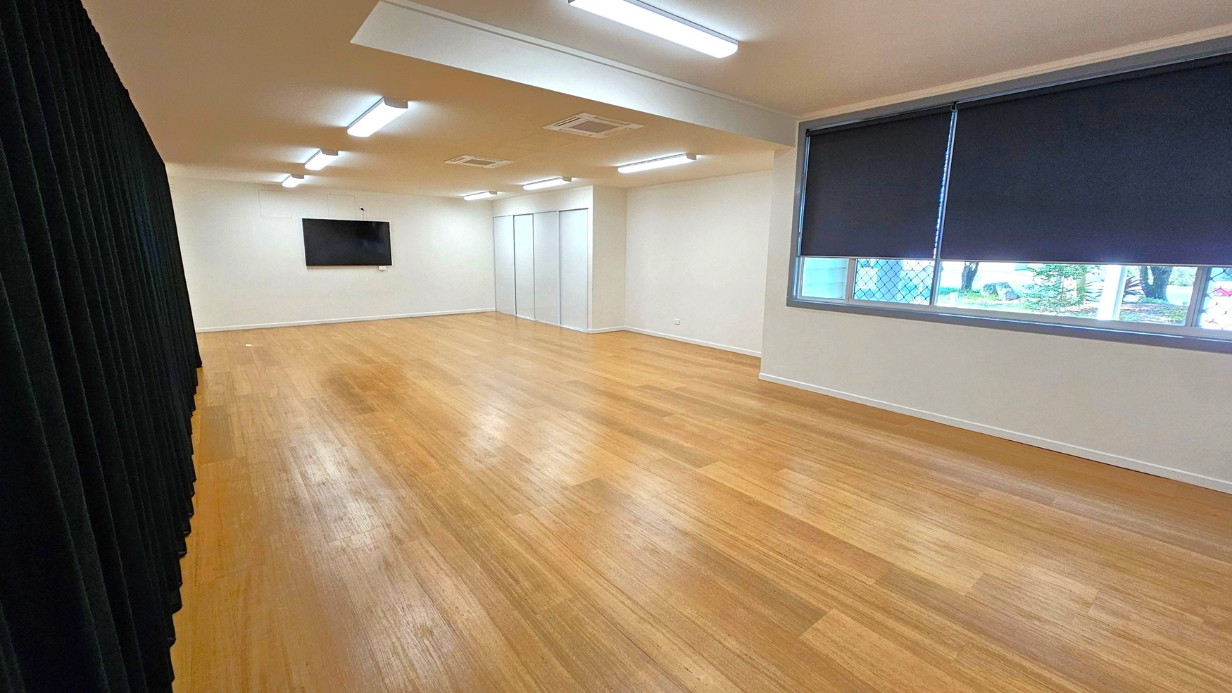 A spacious room with wooden flooring and a white ceiling featuring fluorescent lights. On the left, there is a black curtain. The right side has a large window with dark blinds partially drawn, revealing a view of trees outside. At the far end, a flat-screen TV is mounted on the wall, and there are two white doors. The room has a clean, minimalist appearance.