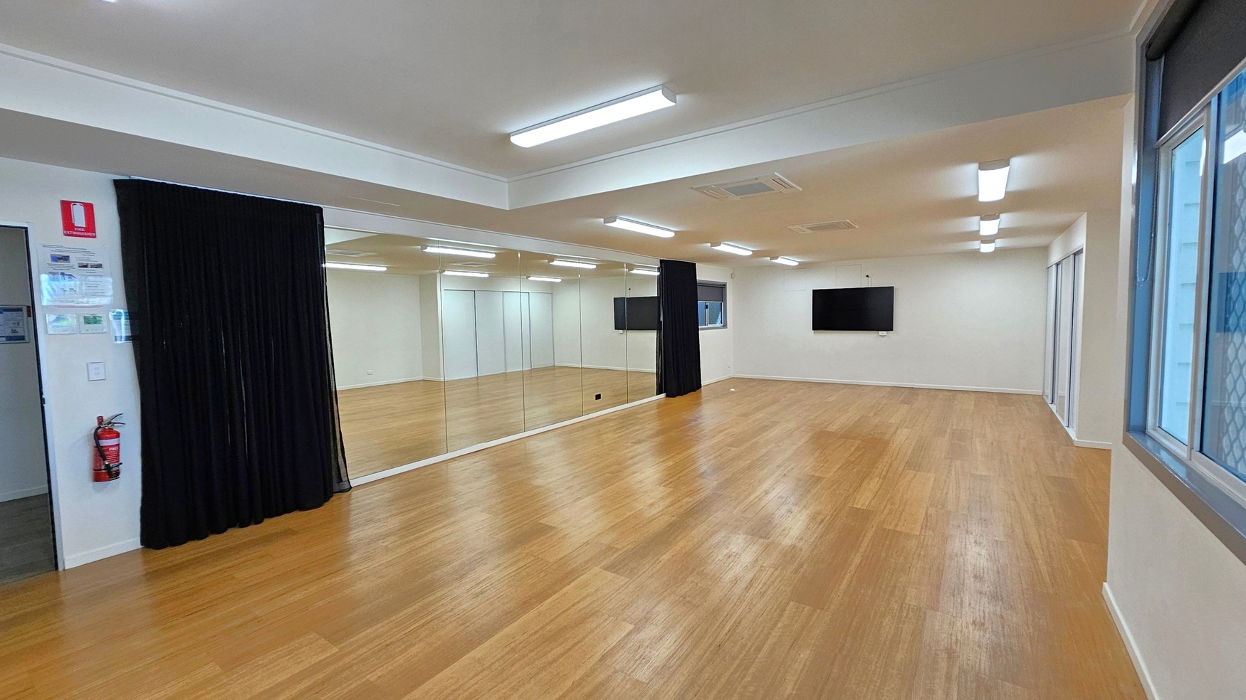 A spacious room with wooden flooring and white walls. On the left, there's a black curtain and a wall with large mirrors. A fire extinguisher is mounted on the wall near the entrance. The right side features a window with a mesh screen and a flat-screen TV mounted on the wall. The ceiling has several fluorescent lights, and the room appears well-lit.