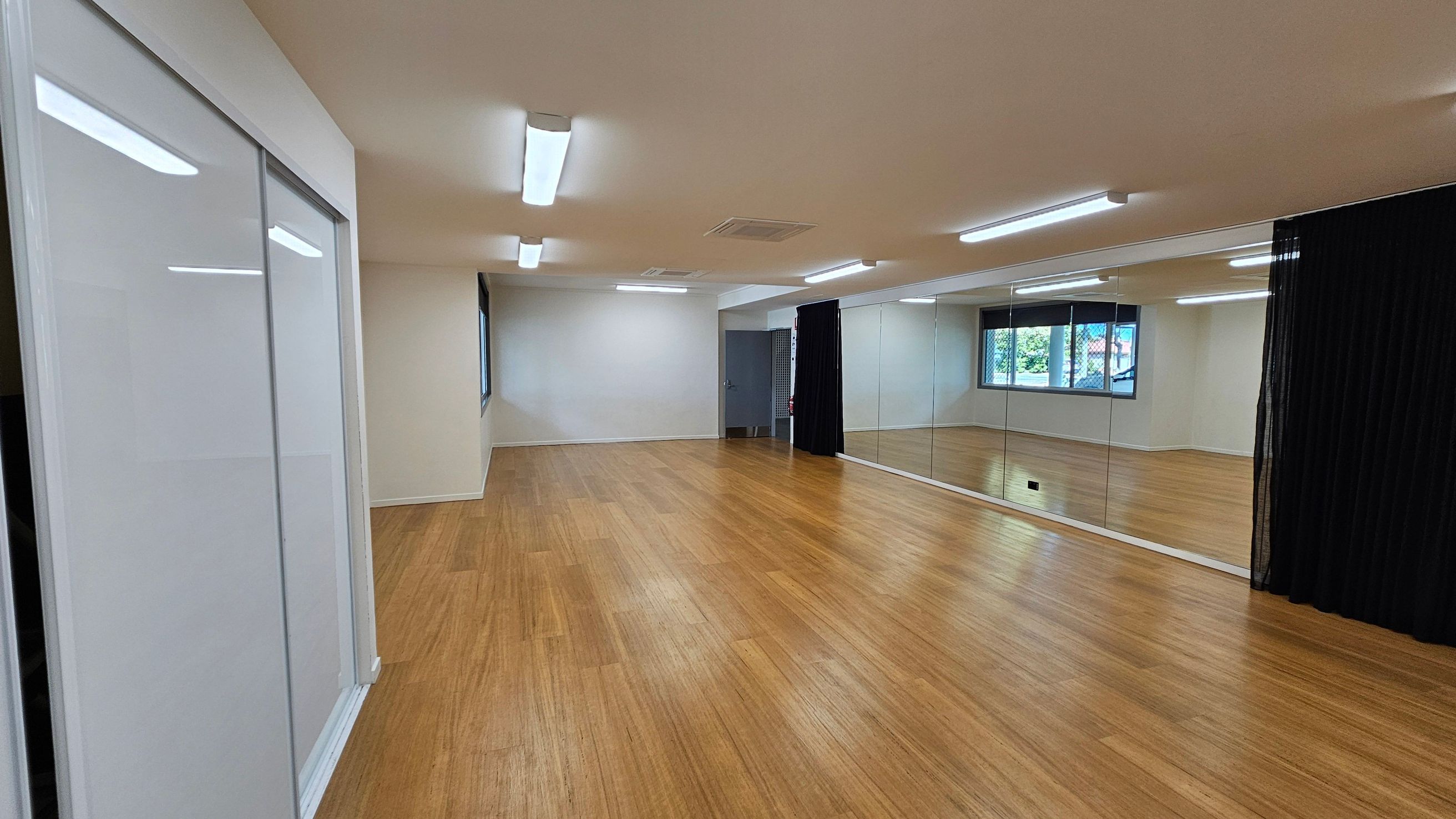 A spacious room with wooden flooring and a white ceiling featuring fluorescent lights. On the right, a wall of mirrors reflects the room, with a black curtain partially covering them. The left side has white sliding doors. At the far end, a window with a grid pattern allows natural light in, and a door is visible next to it. The room appears empty and well-lit.