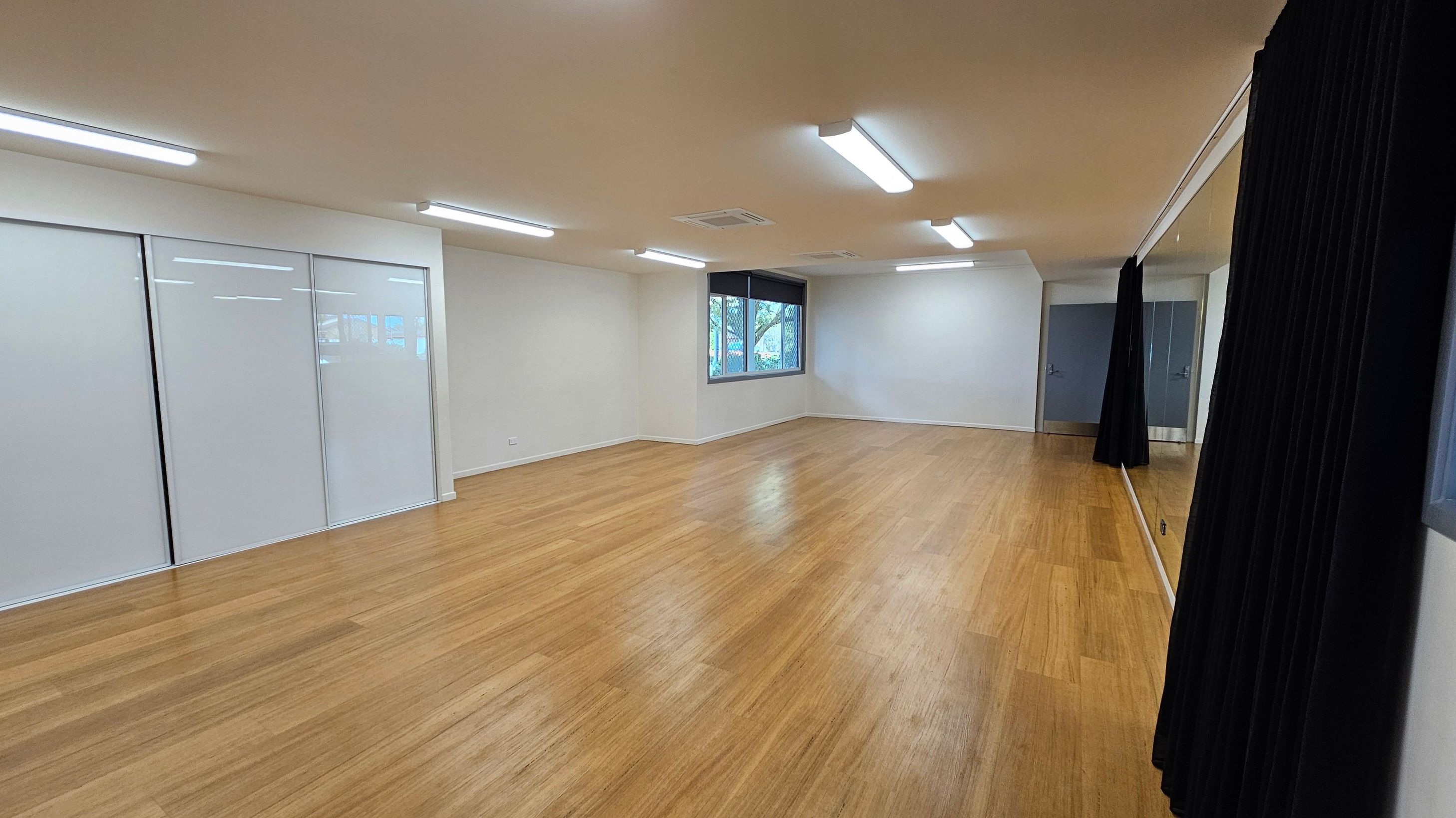 A spacious room with a wooden floor and white ceiling featuring fluorescent lights. On the left, there are large white sliding doors. The right side has a wall-length mirror partially covered by black curtains. At the far end, a window with a black blind overlooks greenery. The room is empty, with light reflecting off the floor.